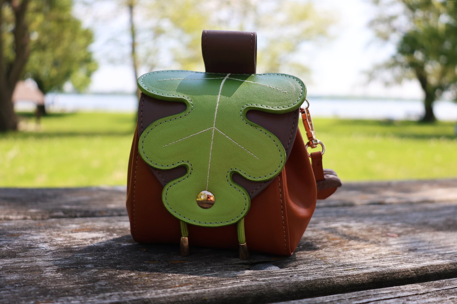 Whimsical forest-inspired pochette featuring a green oak leaf flap and brown faux leather body, photographed outdoors beside a lakeside park.