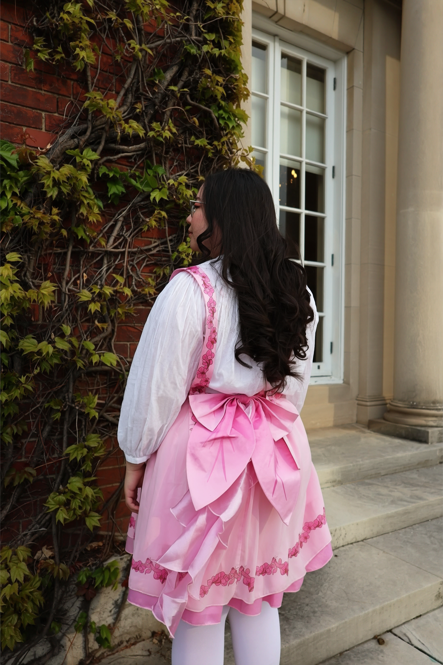 Back view of the Orchid Pinafore Skirt featuring a large pink bow, layered ruffle details, and floral lace trim against an ivy-covered cottage-style wall.