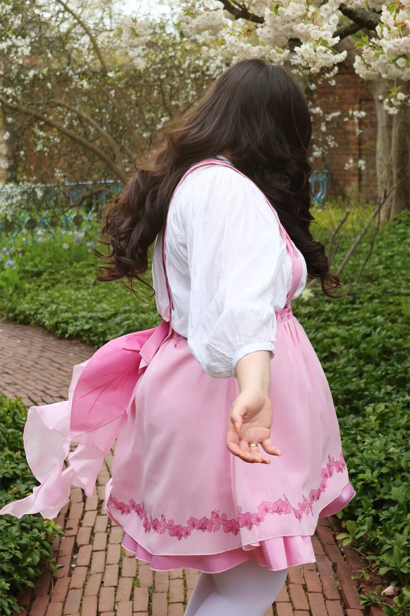 Model wearing the Orchid Pinafore Skirt, a pink fairycore botanical pinafore dress with flowing ribbon details, walking through a flower garden pathway.
