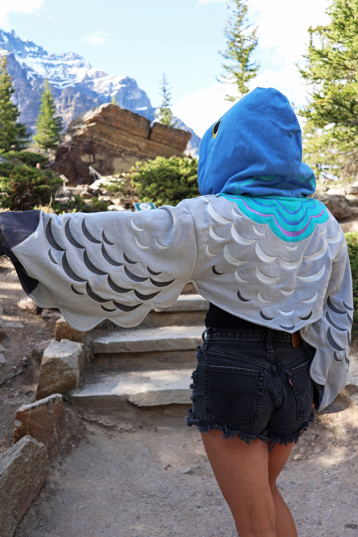 Back view of the Pigeon Hoodie Shrug showcasing layered embroidered feather details across the sleeves and back, blue pigeon hood, and cropped oversized fit in a mountain setting.