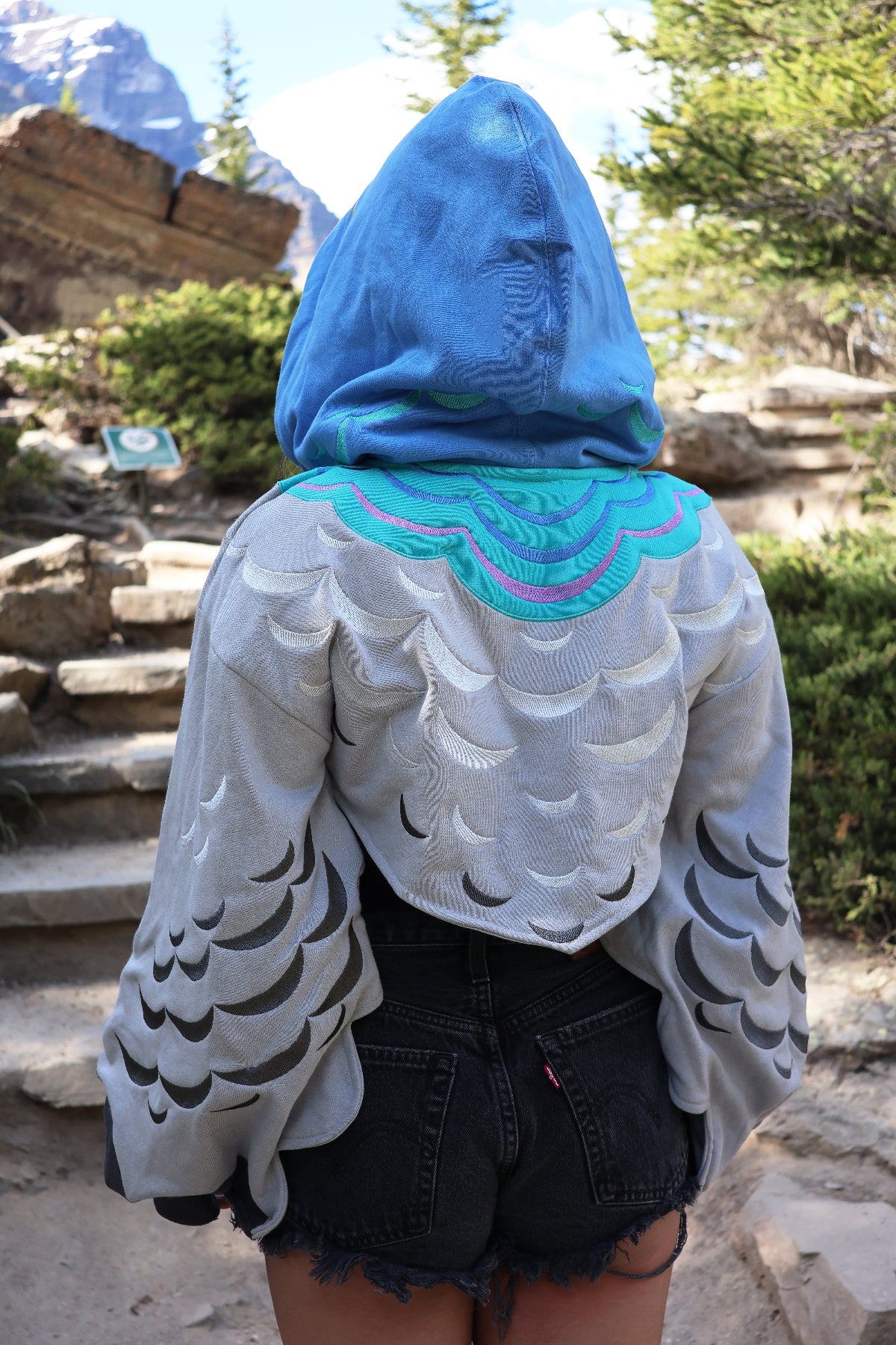 Back view of the Pigeon Hoodie Shrug featuring embroidered feather details, layered wing sleeves, and a blue pigeon-inspired hood, photographed outdoors on a mountain trail.