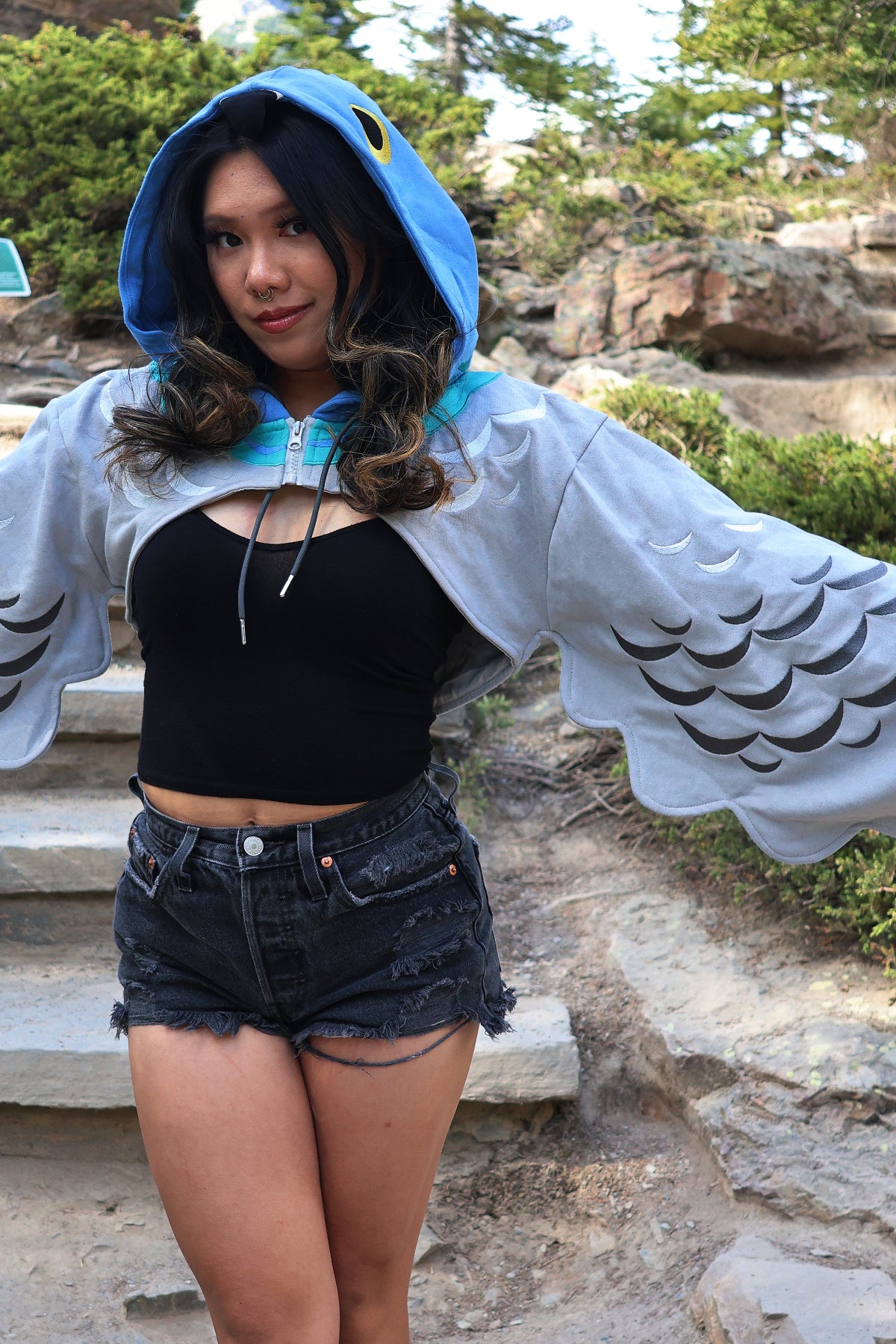 Model wearing the Nyahallo Pigeon Hoodie Shrug with a pigeon-inspired hood, embroidered feather details, and oversized wing-shaped sleeves spread open to showcase the design.