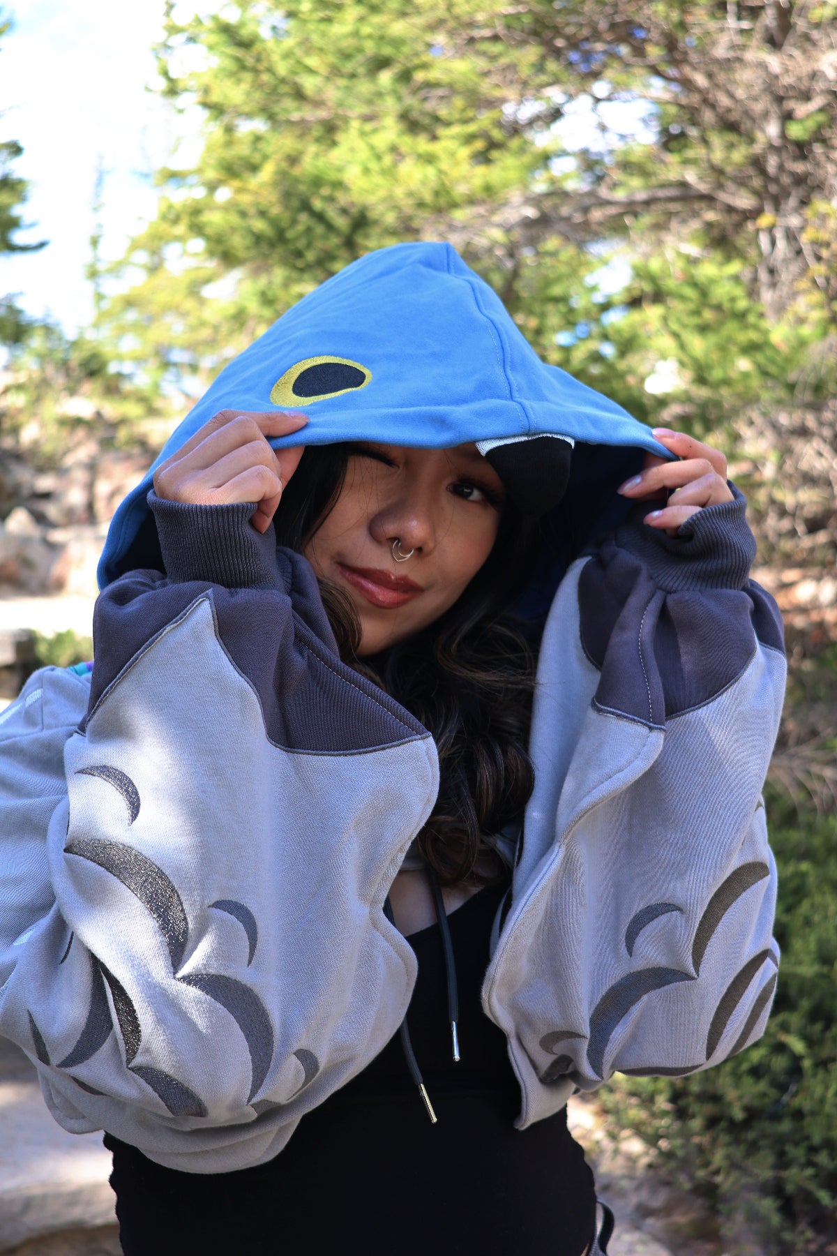 Close-up of the Pigeon Hoodie Shrug featuring a blue pigeon-inspired hood, embroidered feather sleeves, and oversized cropped silhouette, photographed outdoors in nature.