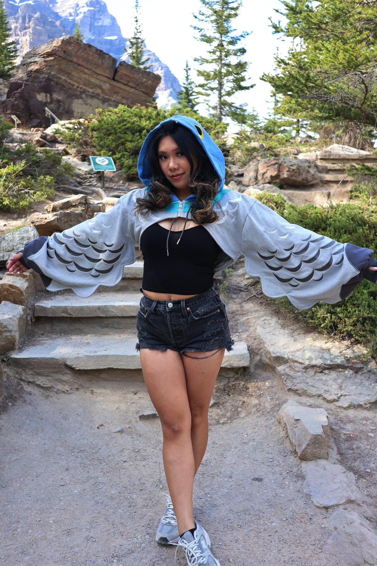 A model wearing the Pigeon Hoodie Shrug outdoors on a mountain trail, featuring a blue pigeon-inspired hood, layered wing sleeves, and feather details with black shorts and a crop top.