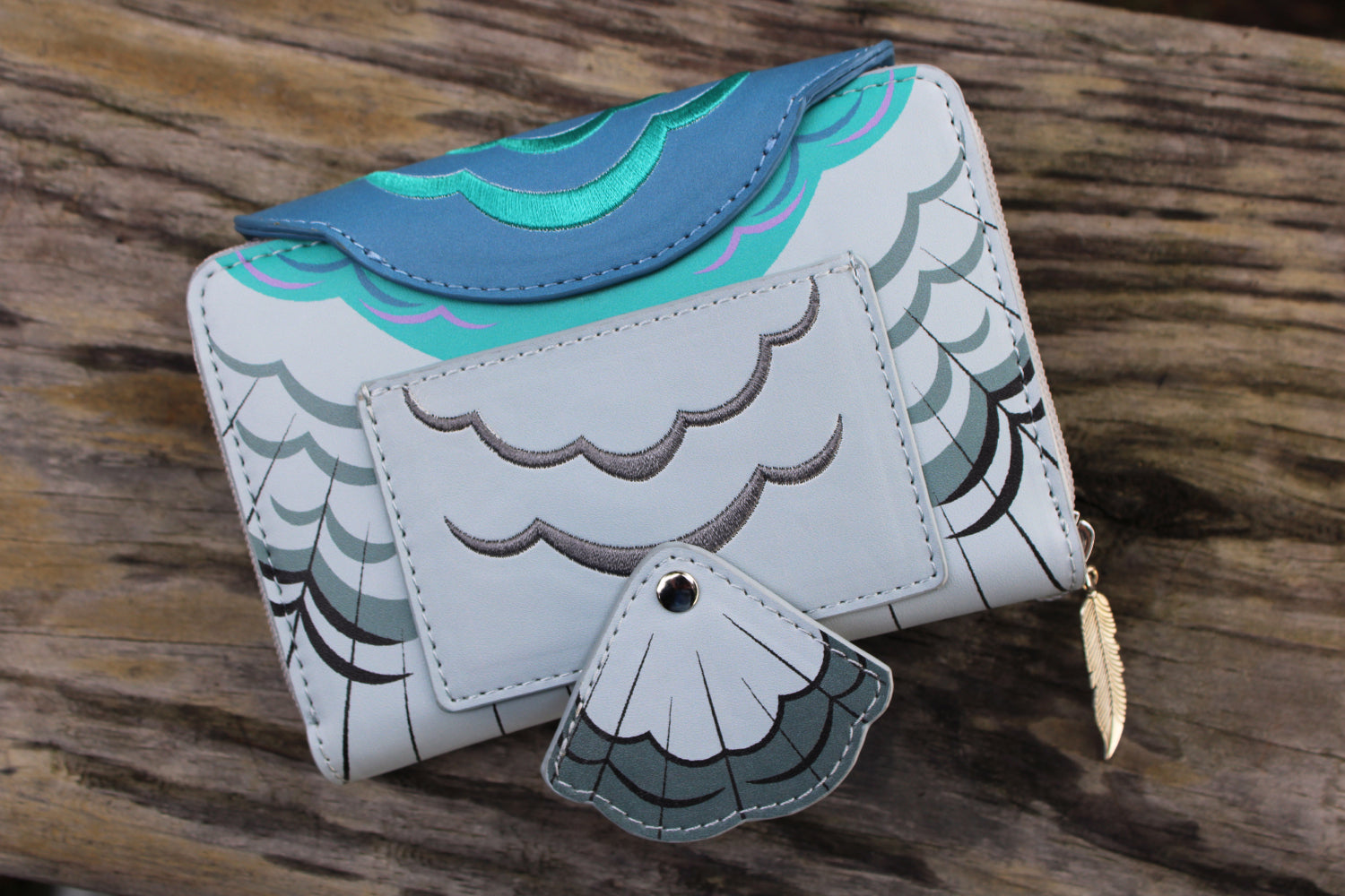 Back view of a whimsical pigeon-shaped vegan leather wallet with layered feather details and a feather zipper charm on a rustic wooden surface.
