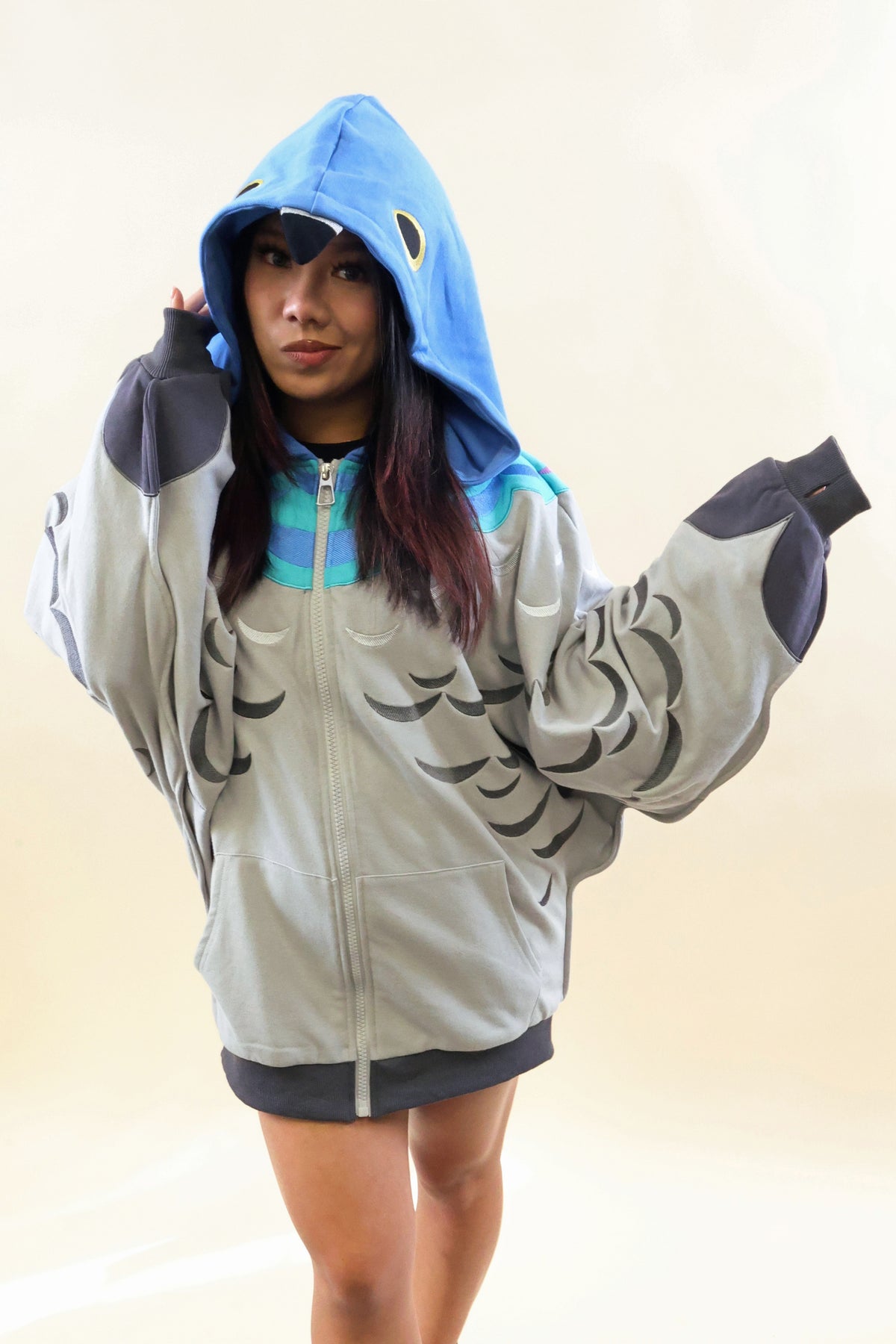 A model wearing an oversized light gray hoodie with a blue shark hood, featuring dark gray pectoral fin graphics on the front and dark gray ribbed cuffs and hem.