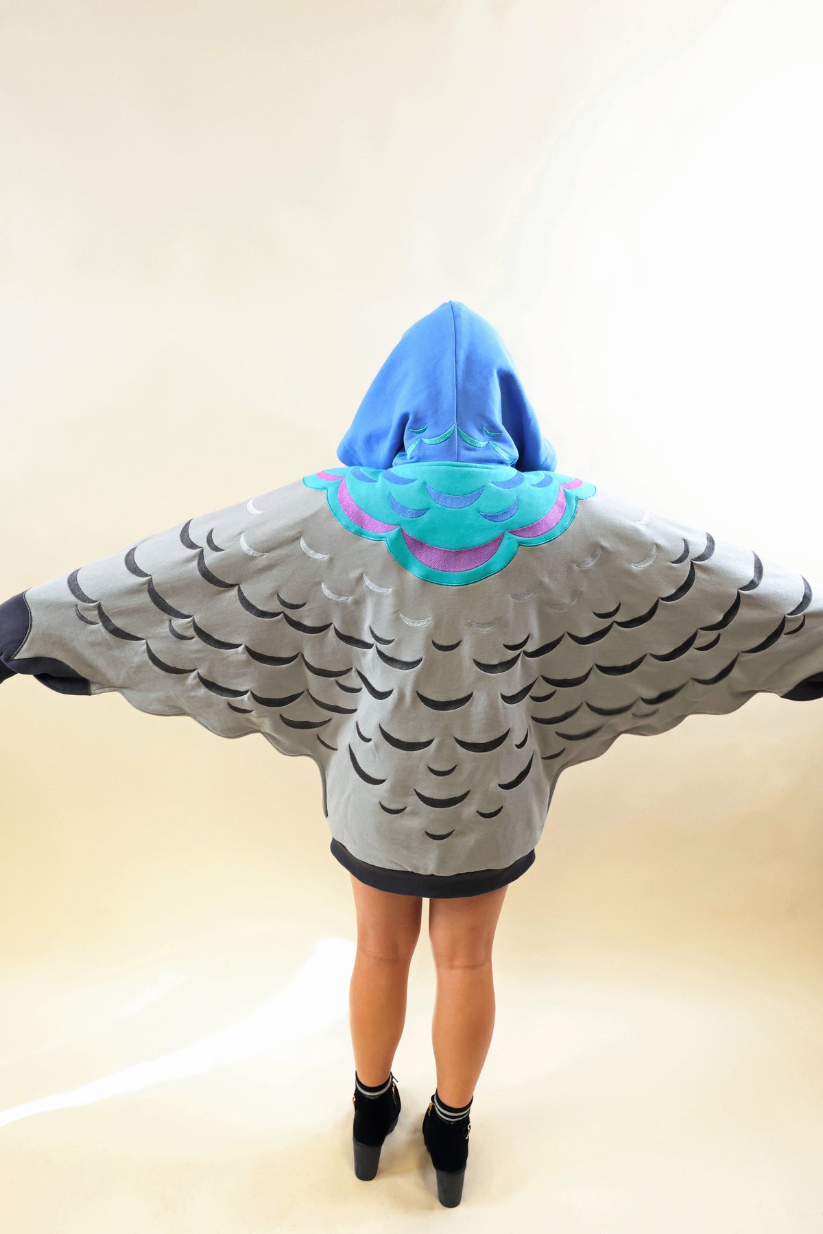 A person viewed from behind wearing a hooded wingsuit-style costume with a blue hood, turquoise and pink collar detail, and a gray body printed with black wing patterns. The person stands with arms outstretched against a plain light beige studio background.