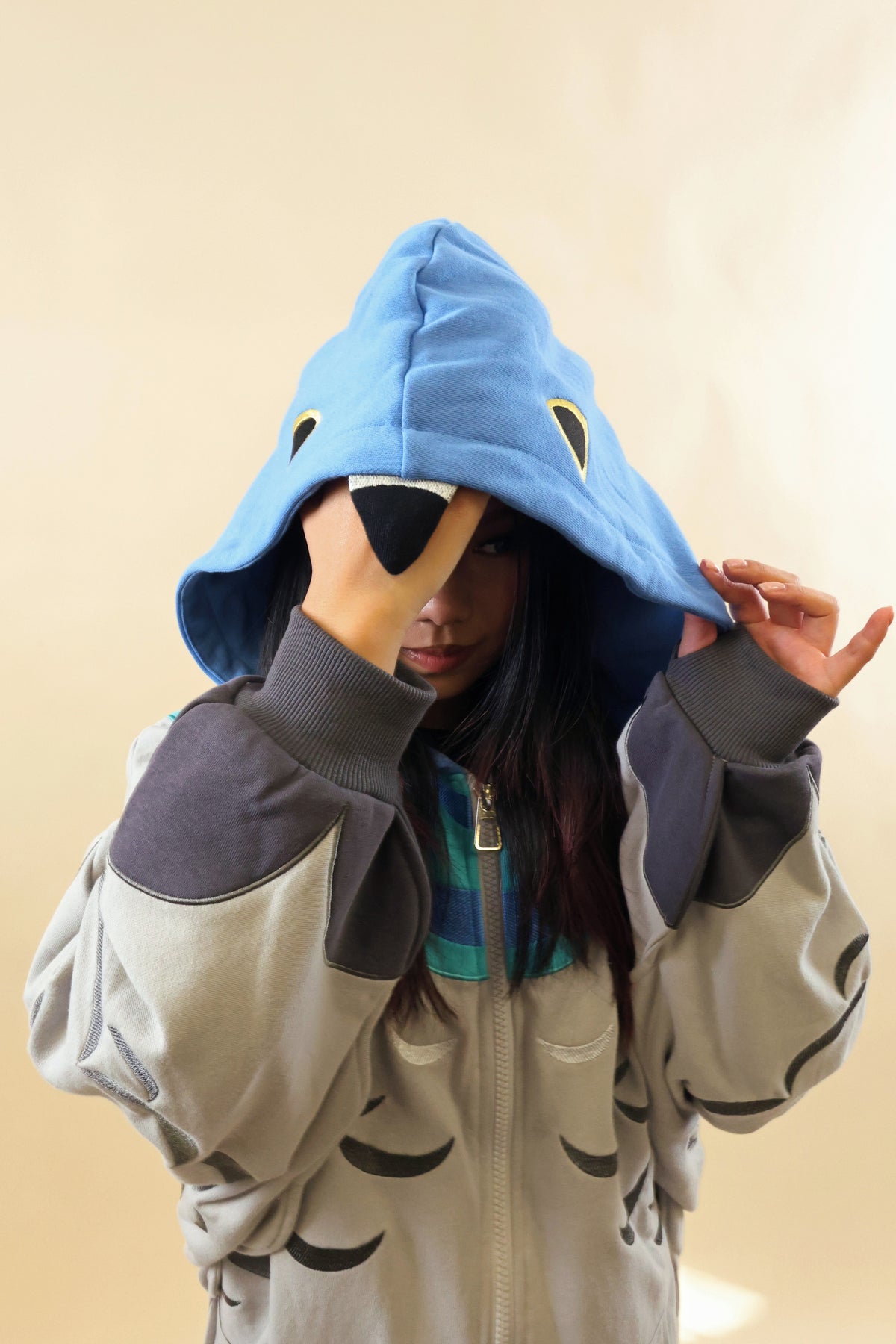 A person wearing a light blue hooded sweatshirt with a shark face design, layered over a gray and black zip-up hoodie, posing against a plain beige background.