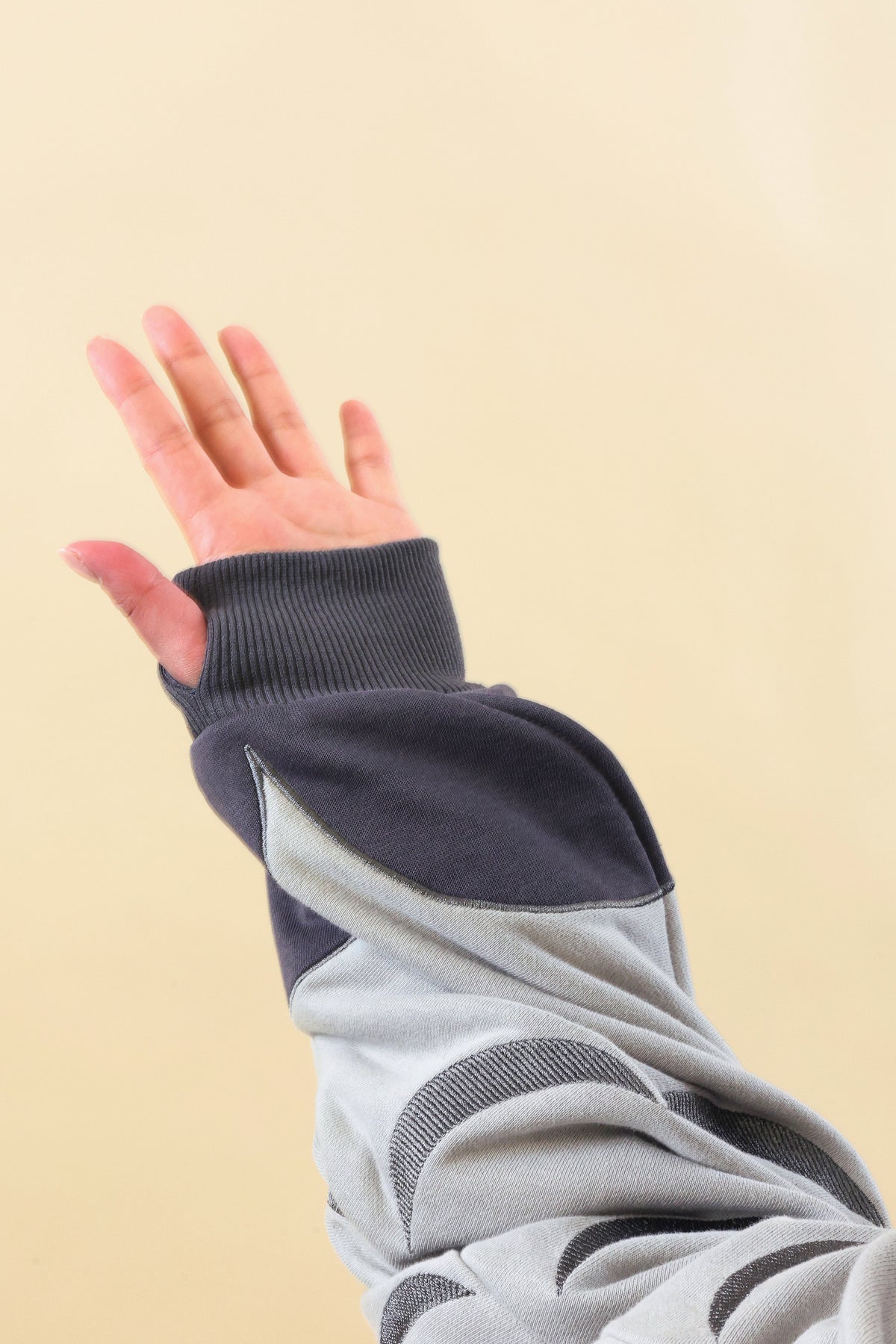 A close-up of a person's raised hand wearing a gray and dark navy long-sleeve top with ribbed cuffs, set against a solid pale beige background.