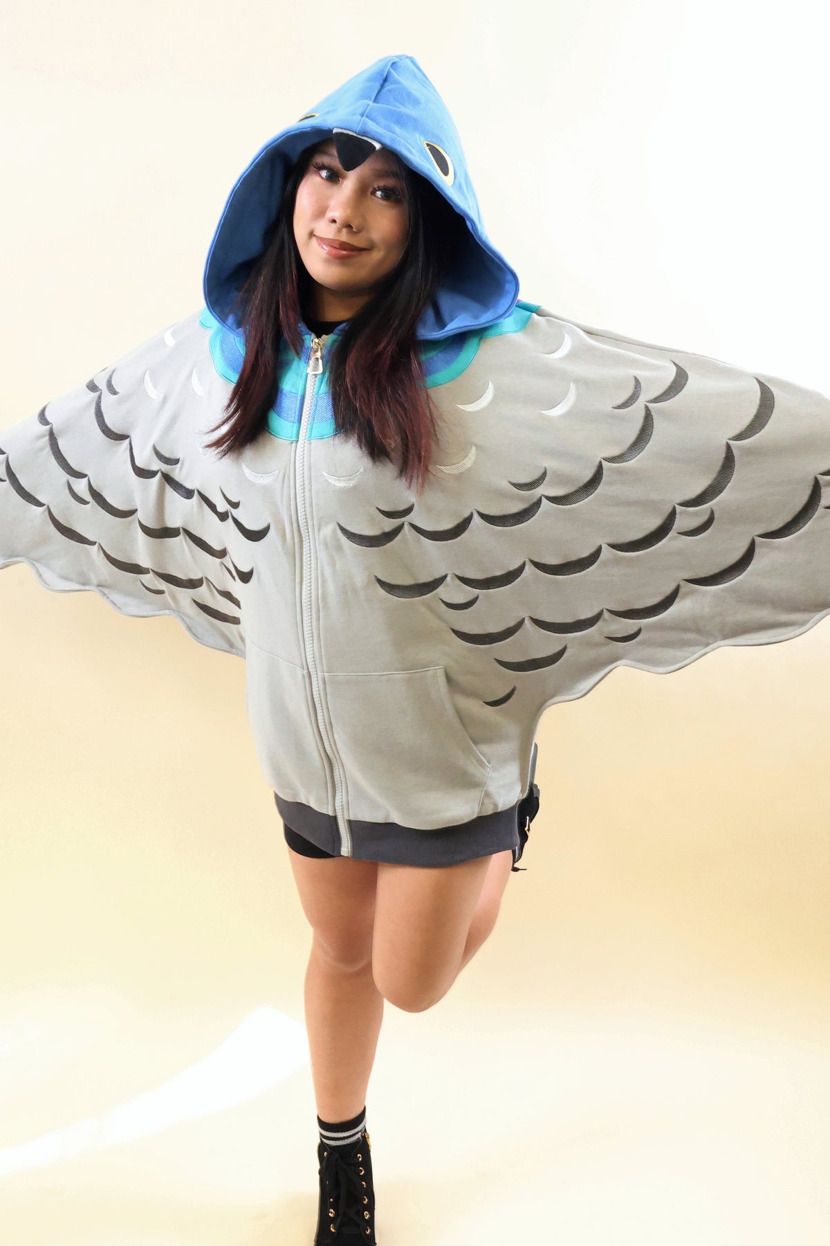 A woman modeling a light gray hooded zip-up sweatshirt designed to look like a bird or fish, featuring large black scalloped wing patterns and a bright blue hood lining.