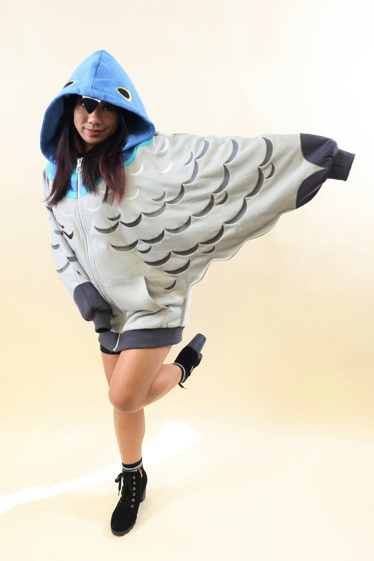 A person modeling a fish-themed hooded costume against a plain beige studio background. The costume is light gray with darker gray scale patterns, a bright blue hood, and dark gray trim, with the model wearing black ankle boots.