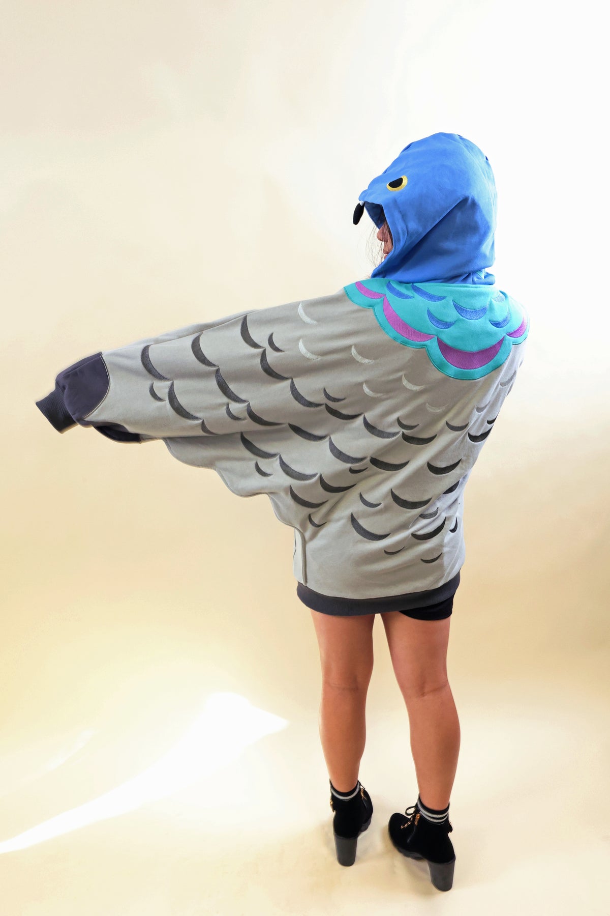 A person shown from the back wearing a hooded cape or hoodie designed like a bird, with a blue hood, black beak detail, gray wings with black feather patterns, and turquoise and purple shoulder accents. The photo is taken against a plain beige studio background.
