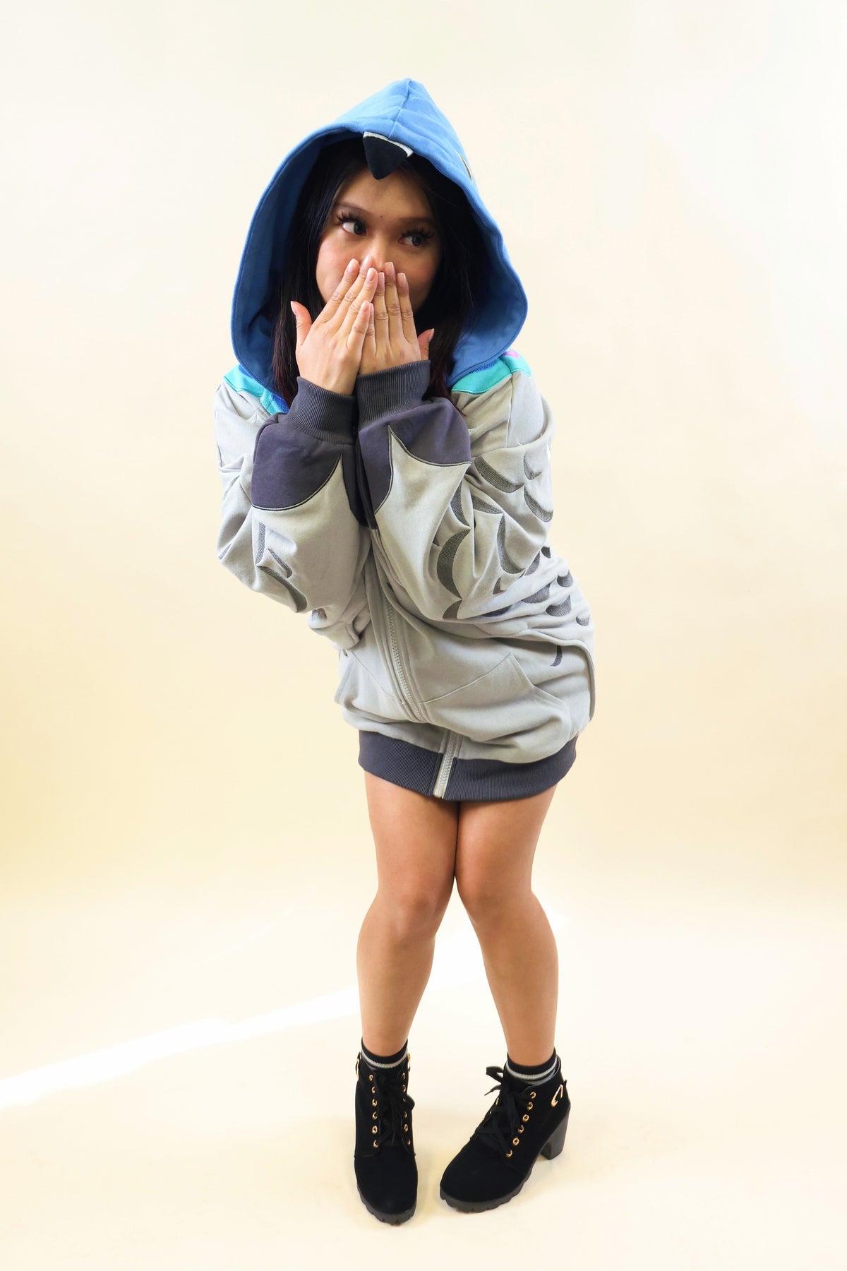 A person wearing a light gray and blue hooded sweatshirt with dark gray shorts and black lace-up ankle boots, standing against a plain beige studio background.