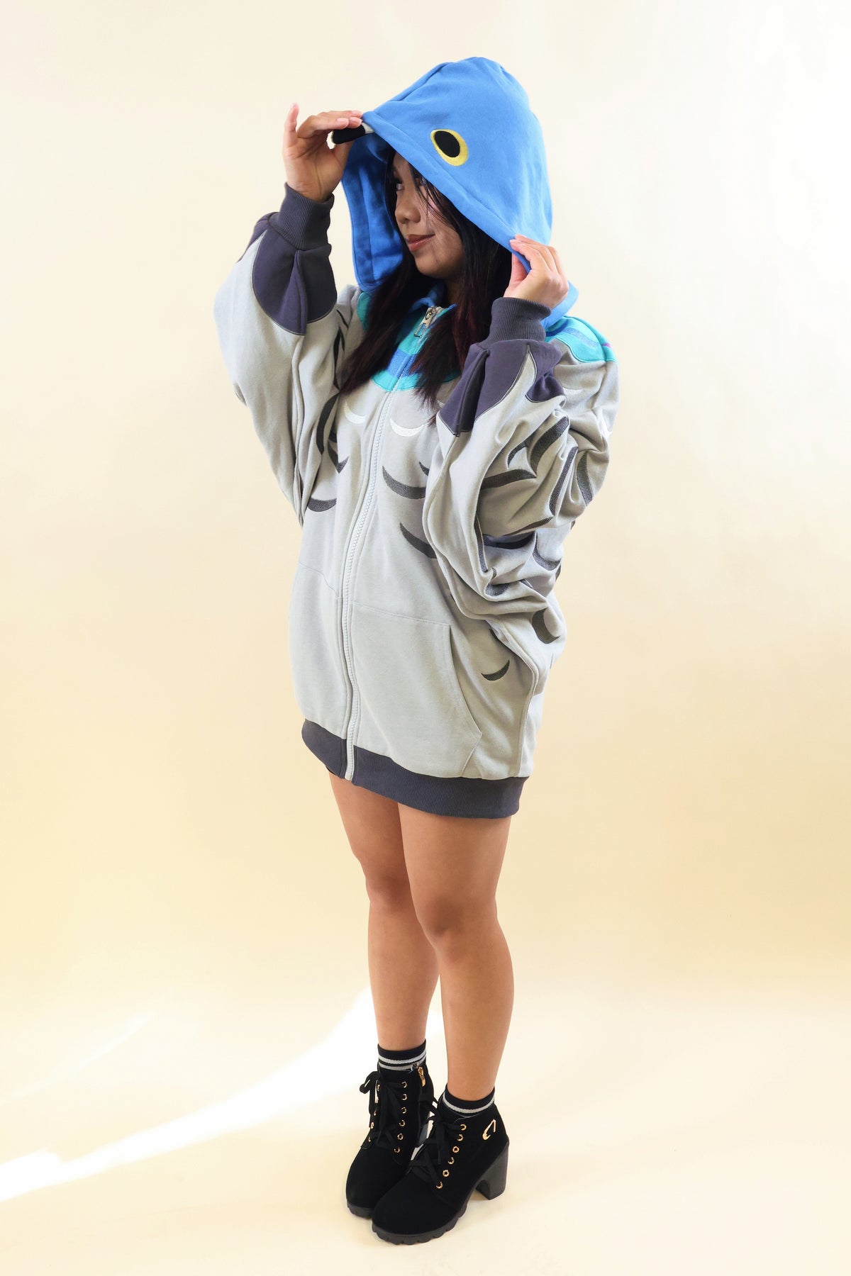 A model wearing a shark-themed zip-up hoodie kigurumi, featuring a light gray body, dark gray details, and a bright blue hood with a yellow eye design, set against a plain beige studio background.