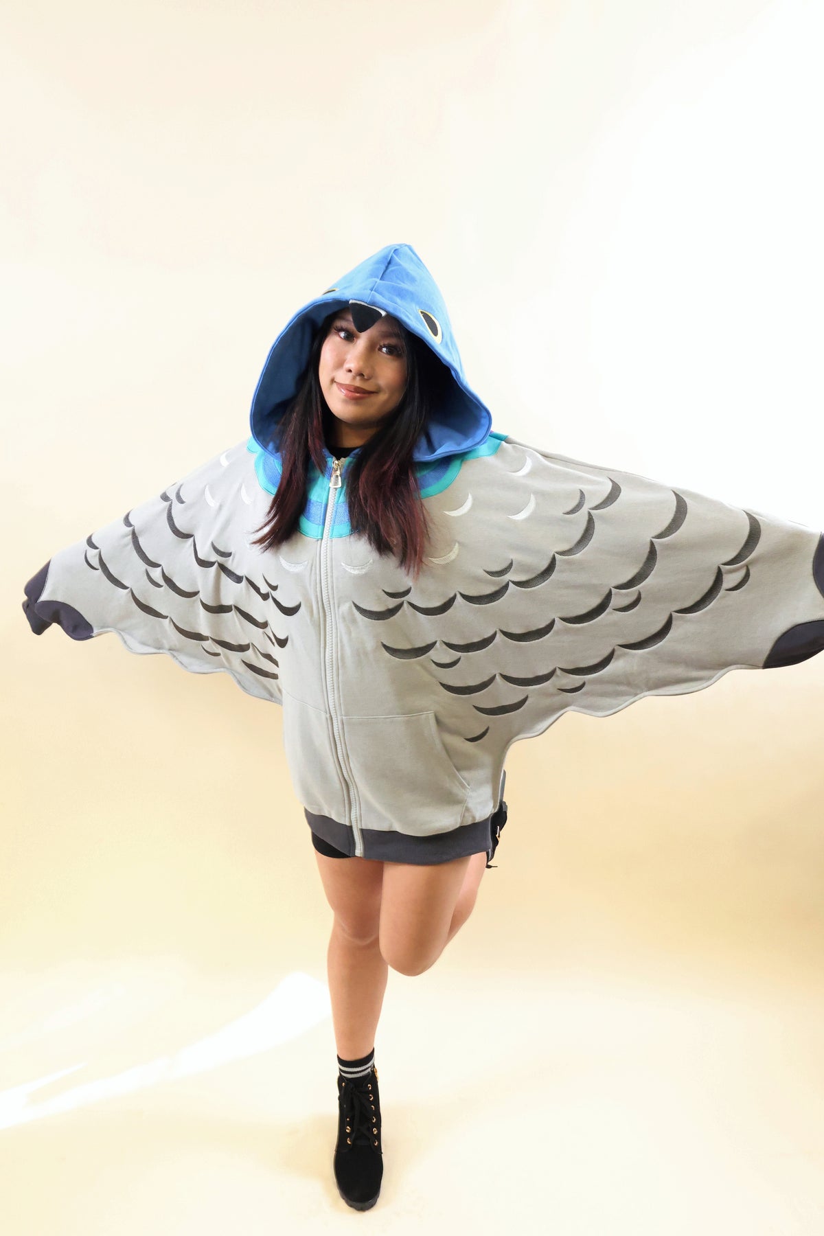 A woman modeling a bird-themed zip-up hooded sweatshirt with wings printed on the front, featuring a light gray body, black wing patterns, and a bright blue hood lining, set against a plain beige studio background.