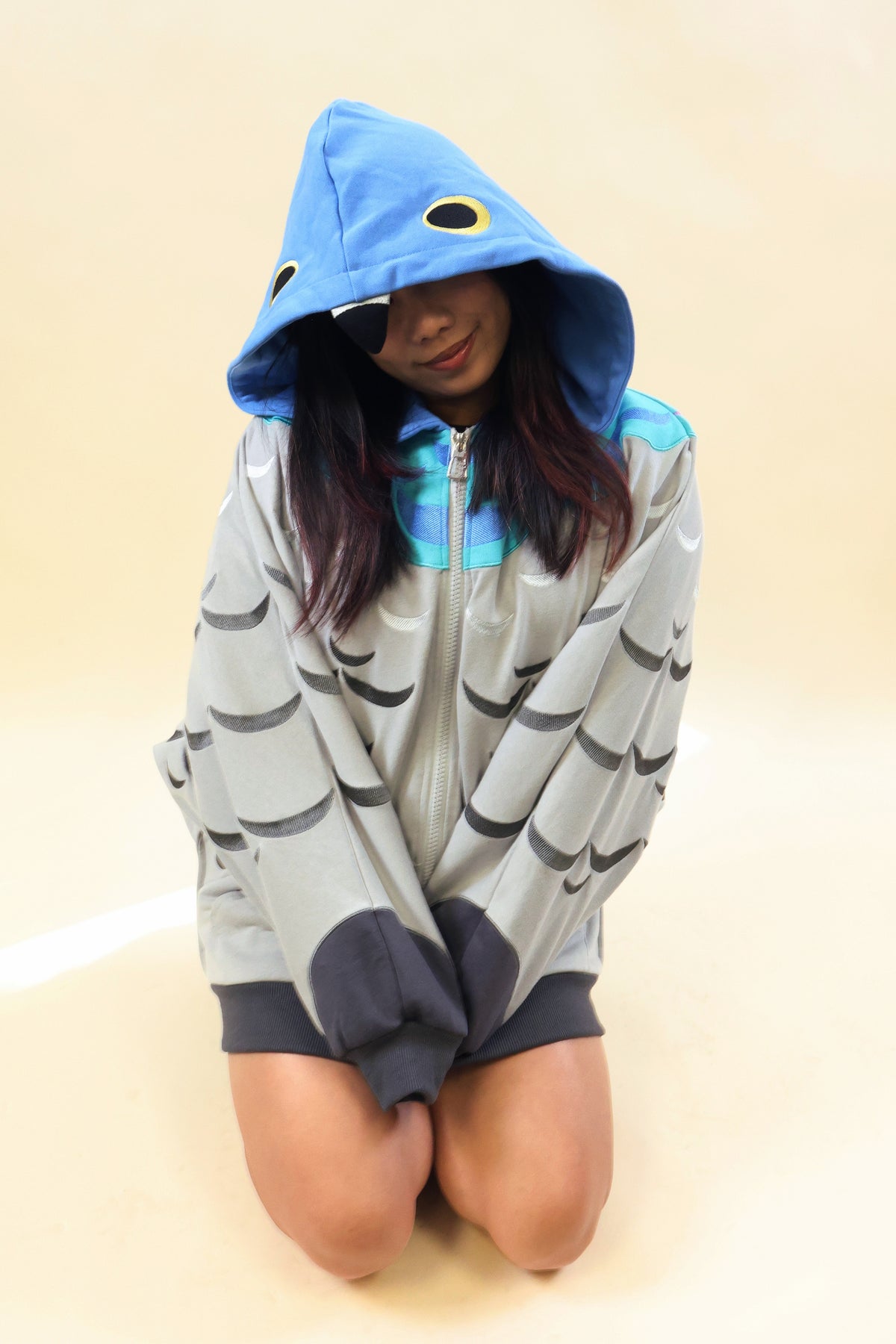 A person kneeling on a plain beige studio background wearing an oversized fish-themed hooded zip-up jacket. The jacket is light gray with dark gray scale stripes, a bright blue hood, and turquoise zipper details.