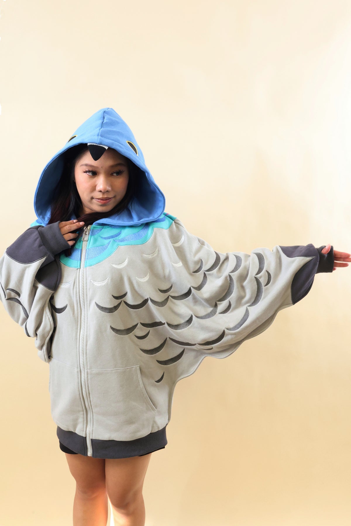 A child wearing a light gray bird-themed hooded zip-up sweatshirt with a blue hood, black wing patterns, and a turquoise chest accent, posed against a plain beige background.