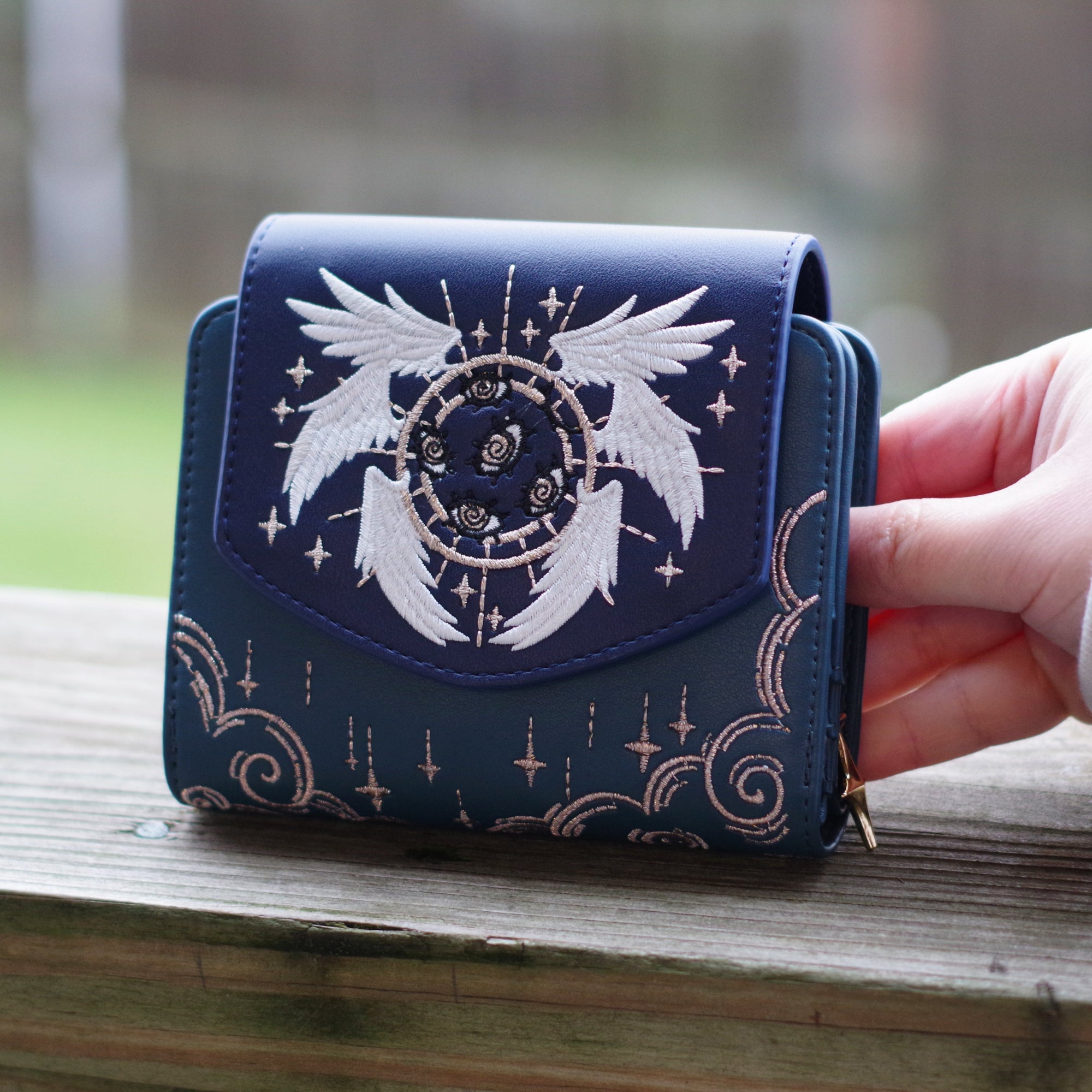 Blue celestial angel wallet with embroidered white wings, star details, and gold accents resting on a wooden railing outdoors.