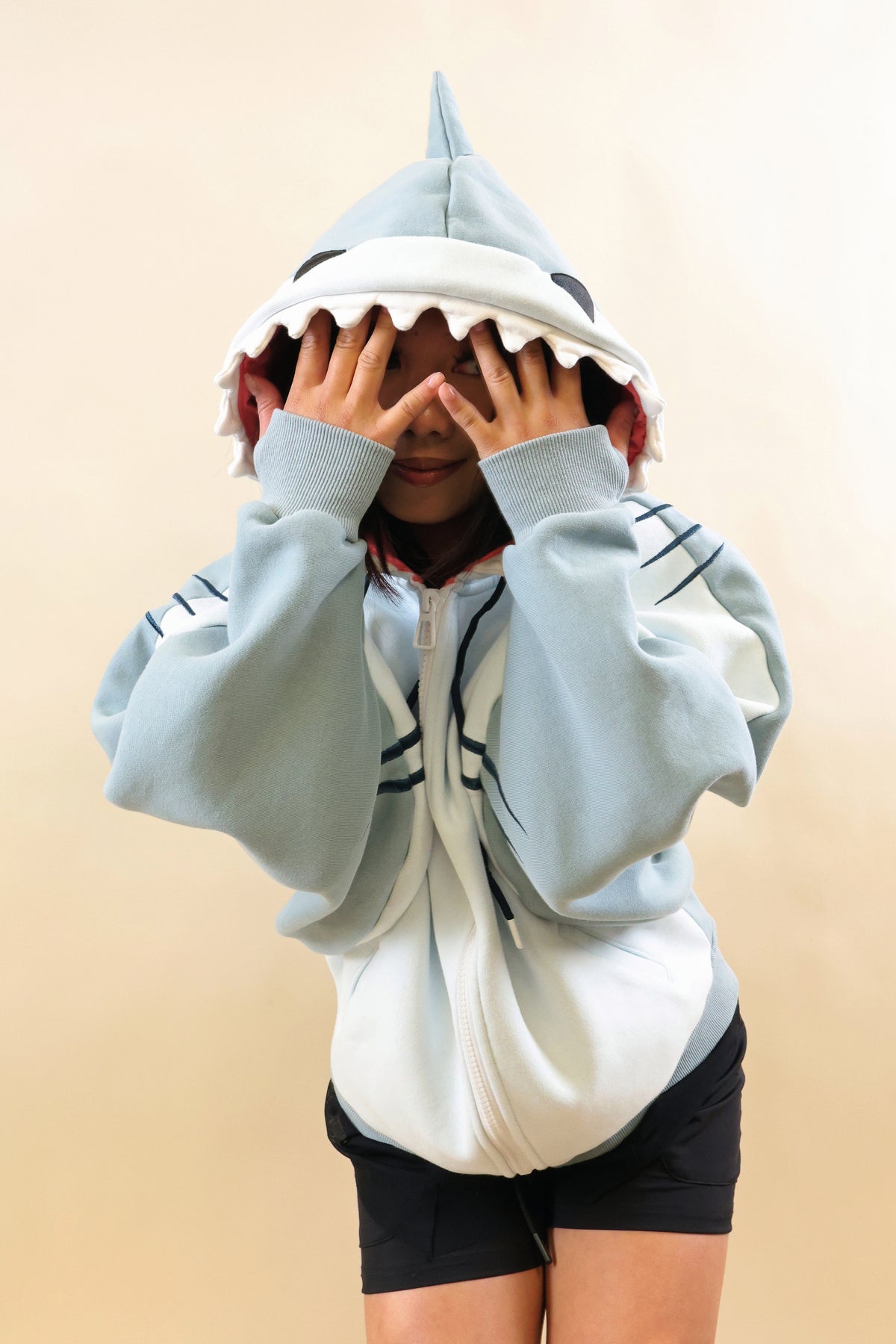 Model wearing the Nyahallo Shark Hoodie, featuring a light blue and white shark design with a dorsal fin, tooth-lined hood, embroidered gill details, and oversized sleeves, playfully covering her eyes.