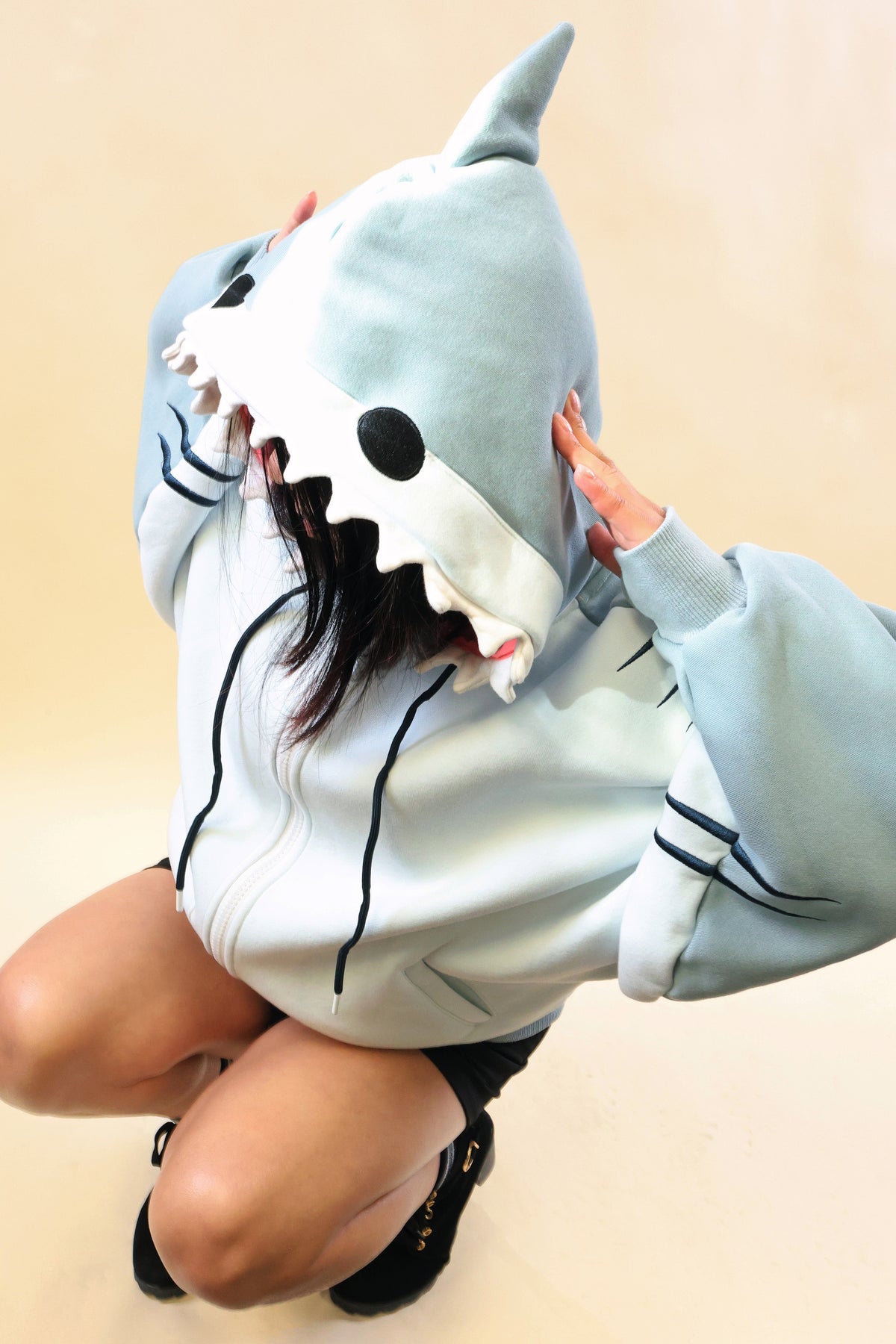 Model kneeling in the Nyahallo Shark Hoodie, showcasing its light blue shark hood with dorsal fin, black eyes, white teeth, oversized sleeves, and embroidered gill details.