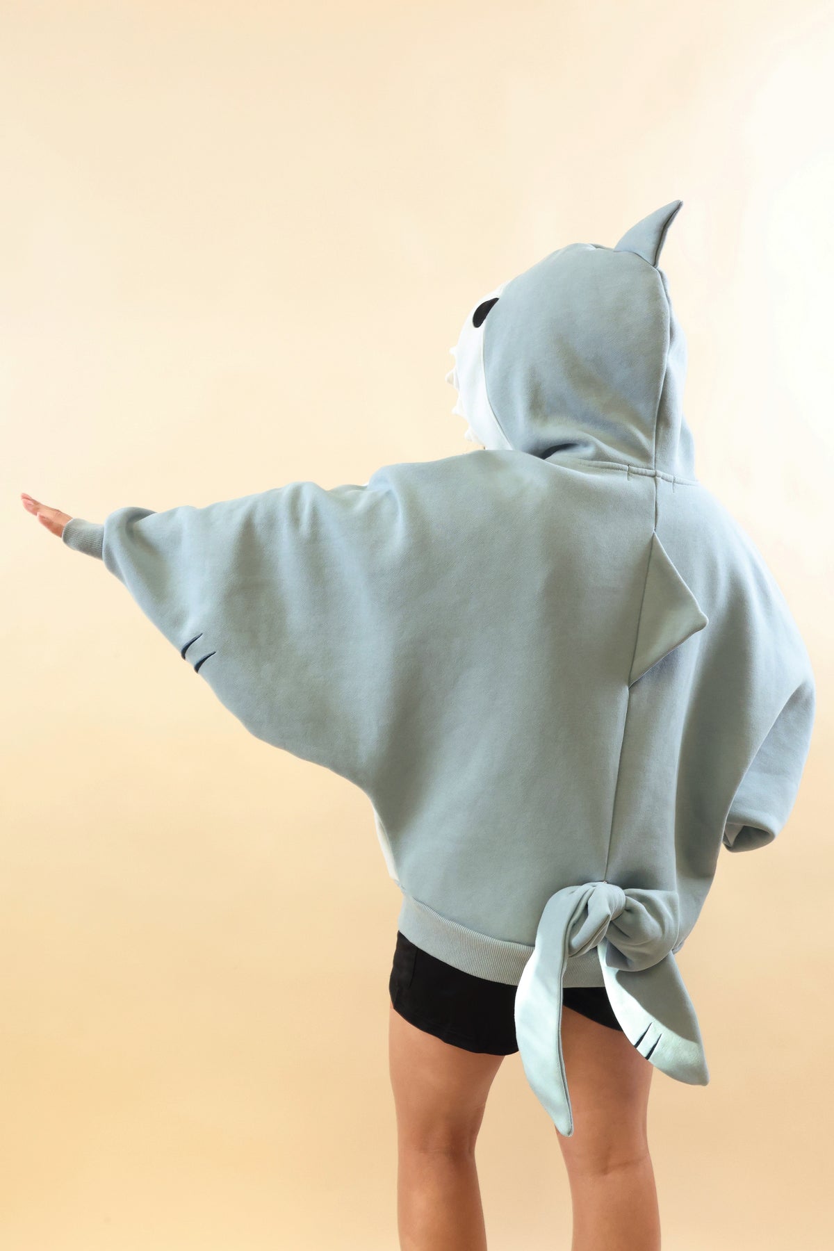 Back view of a model wearing the Nyahallo Shark Hoodie with one arm outstretched, showcasing its oversized light blue design, shark hood with dorsal fin, back fin, and dangling shark tail.