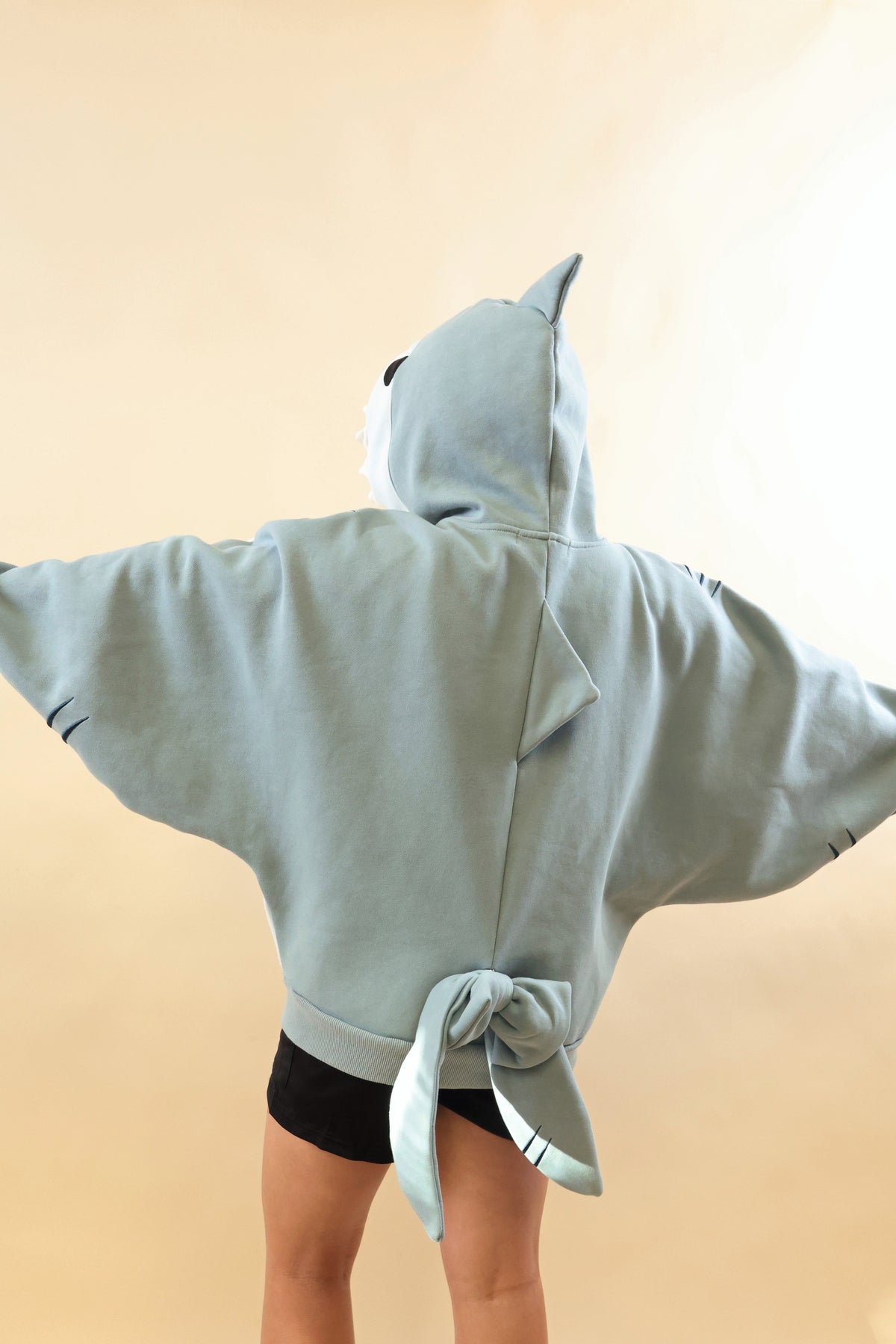Back view of a model wearing the Nyahallo Shark Hoodie with arms outstretched, showcasing its oversized light blue design, dorsal fins, wide sleeves, and dangling shark tail.