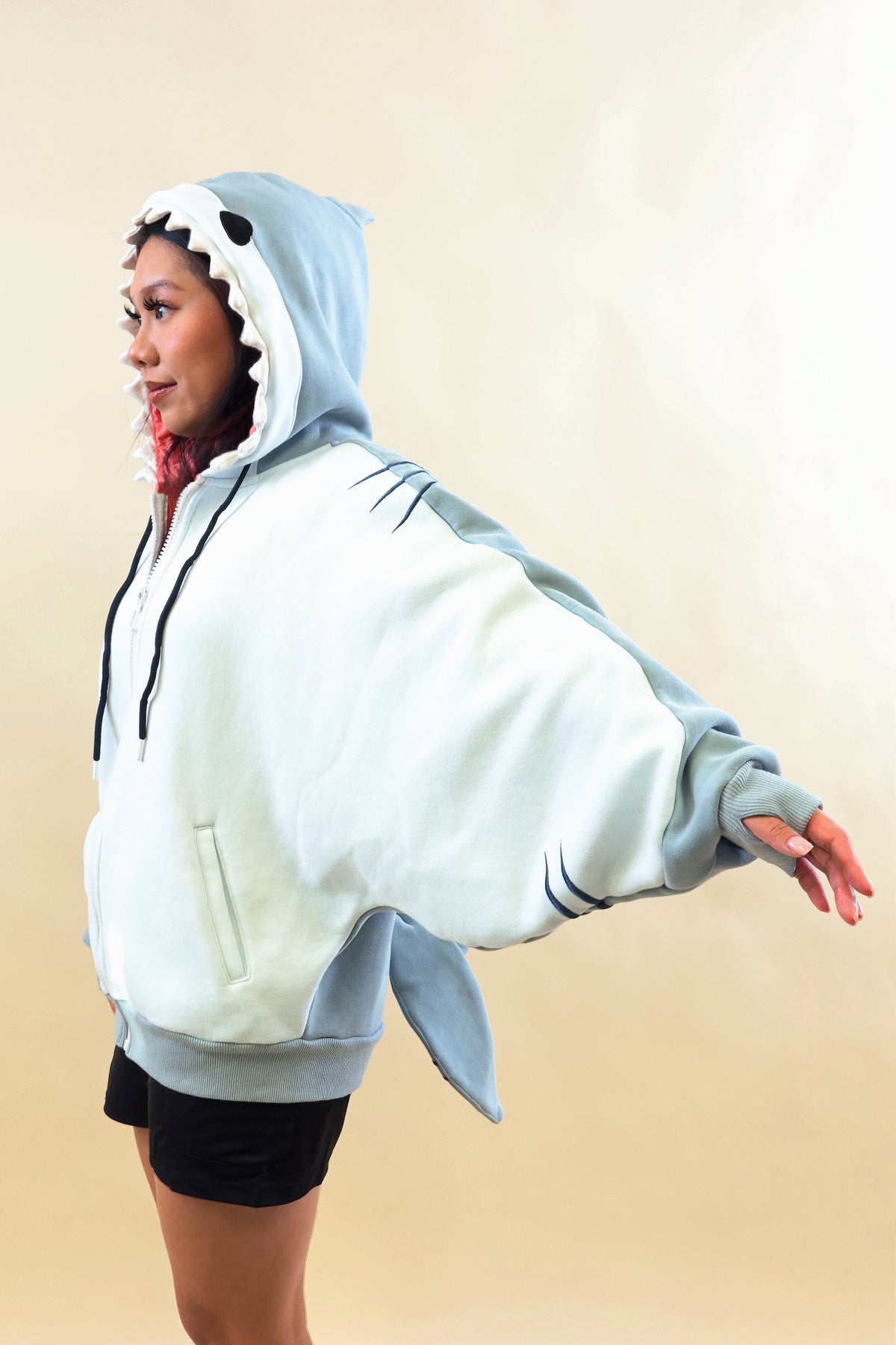Side view of a model wearing the Nyahallo Shark Hoodie with one arm outstretched, showcasing its oversized light blue and white design, shark teeth hood, embroidered gill details, front pocket, and dangling shark tail.