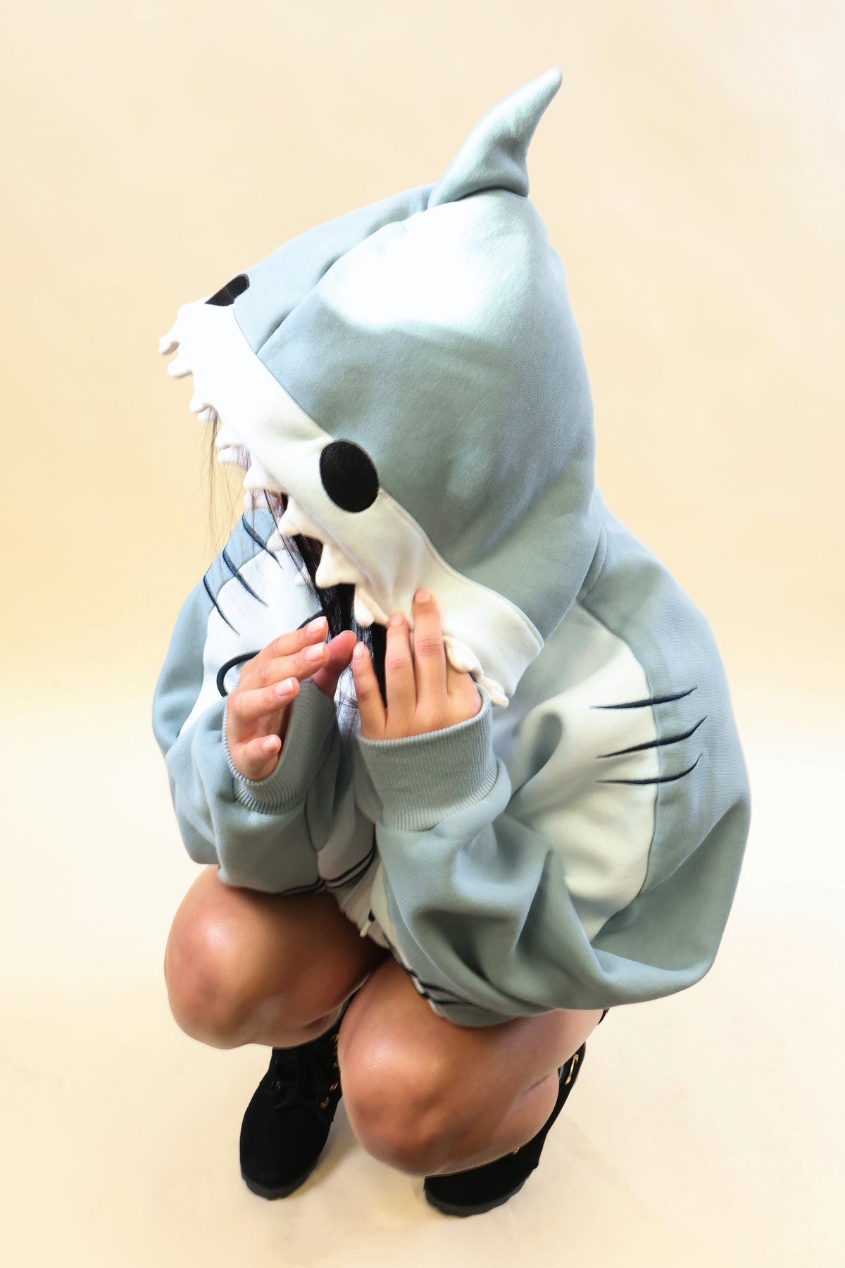 Model crouching in the Nyahallo Shark Hoodie, showcasing its light blue shark hood with dorsal fin, black eyes, white teeth, and oversized sleeves with embroidered gill details.