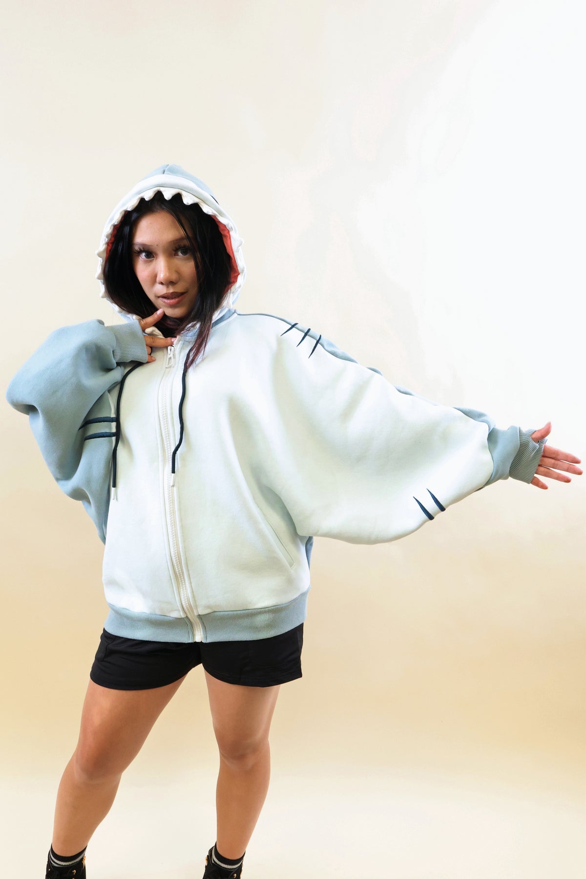 Model wearing the Nyahallo Shark Hoodie with one arm outstretched, showcasing its oversized light blue and white zip-up design, shark teeth hood, embroidered gill details, and wide sleeves.