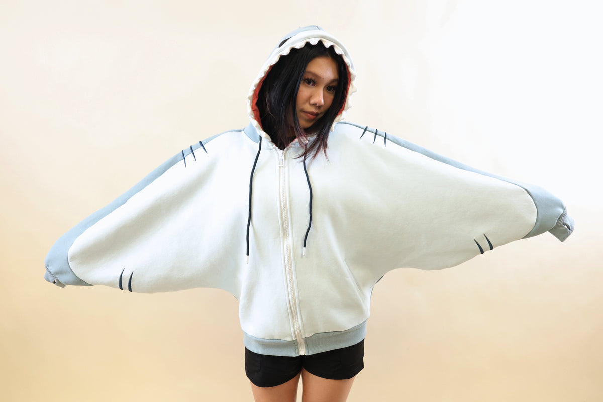 Front view of a model wearing the Nyahallo Shark Hoodie with arms outstretched, showcasing its oversized light blue and white batwing sleeves, shark teeth hood, embroidered gill details, and zip-up design.