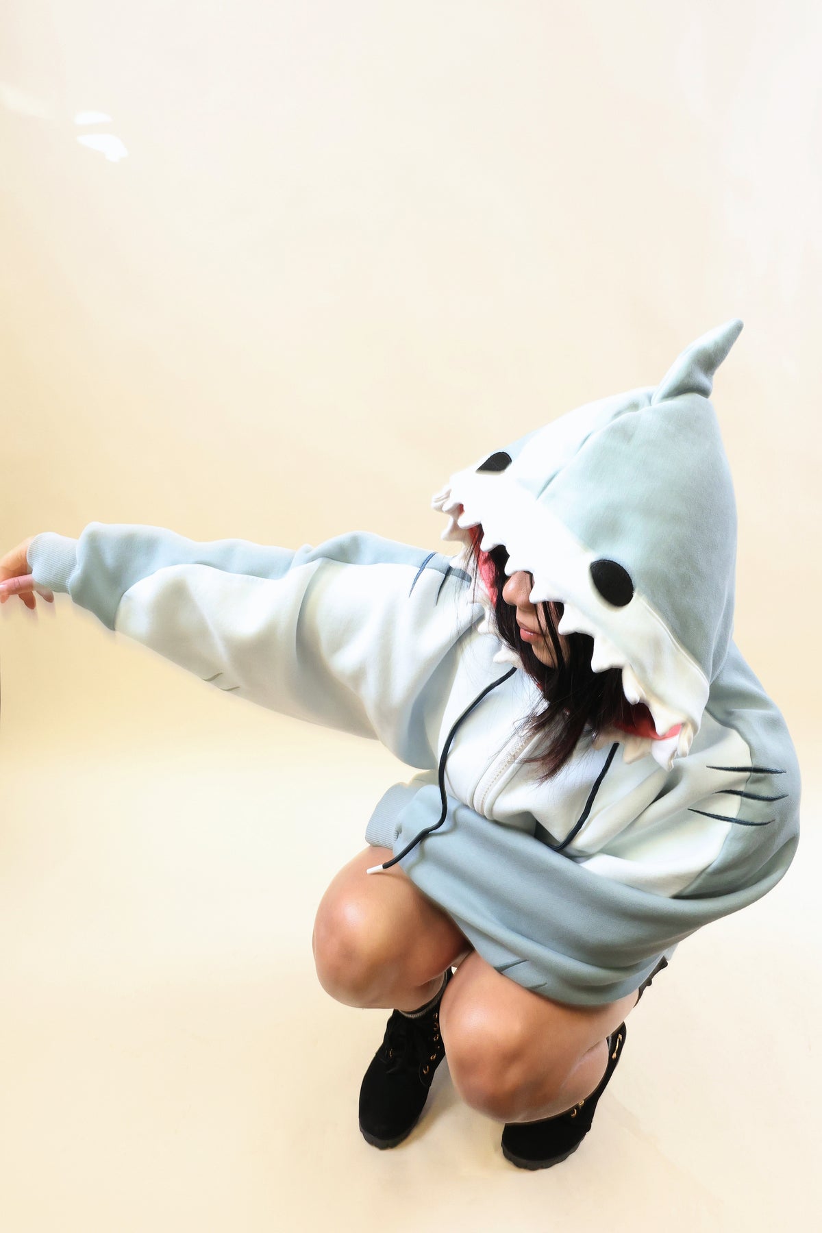 Model crouching in the Nyahallo Shark Hoodie with one arm outstretched, showcasing its light blue shark hood with white teeth, dorsal fin, oversized sleeves, and embroidered gill details.