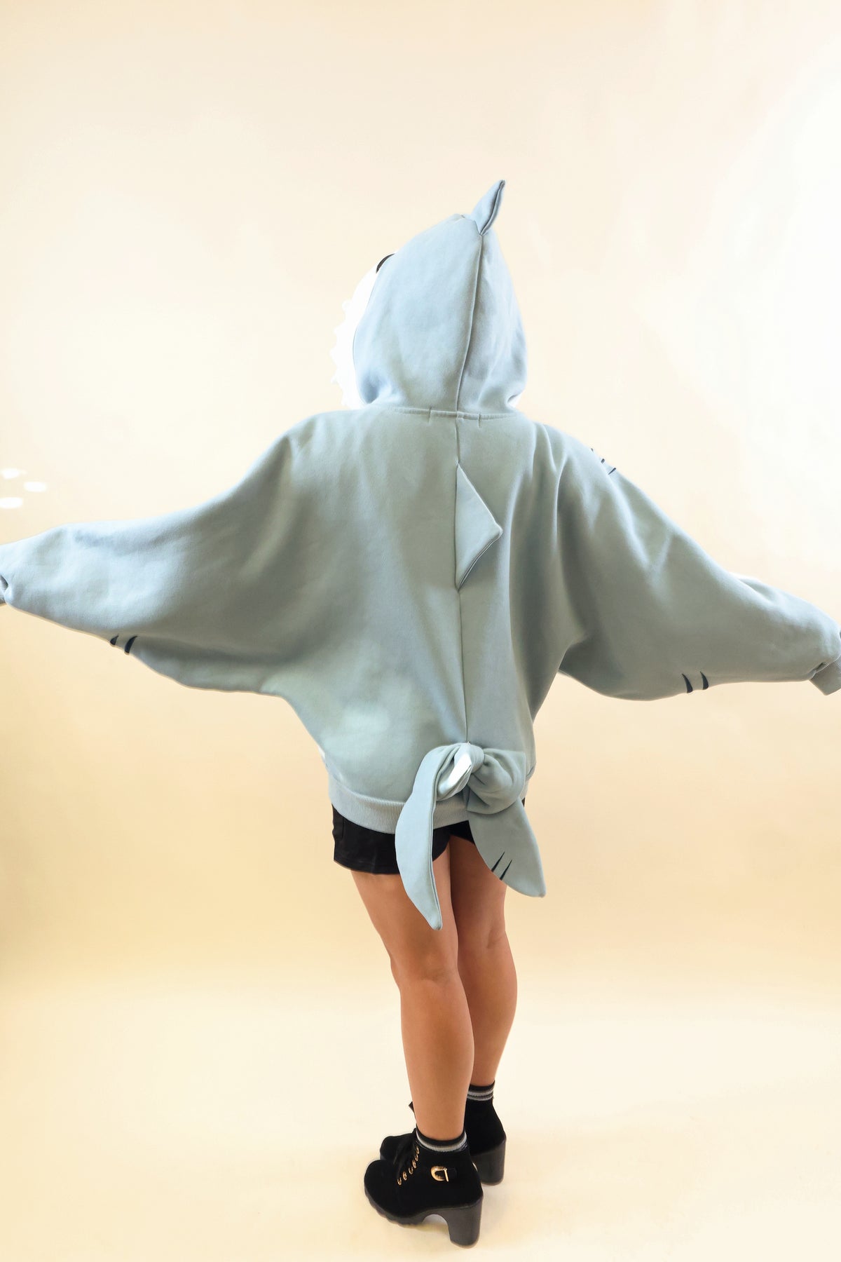 Back view of a model wearing the Nyahallo Shark Hoodie with arms outstretched, showcasing its oversized light blue design, dorsal fins, embroidered gill details, and dangling shark tail.