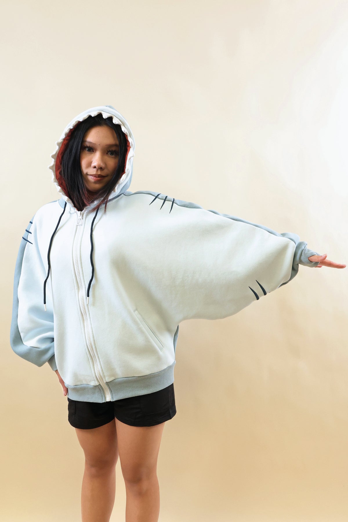 Front view of a model wearing the Nyahallo Shark Hoodie with one arm outstretched, showcasing its oversized light blue and white zip-up design, shark teeth hood, wide batwing sleeves, and embroidered gill details.