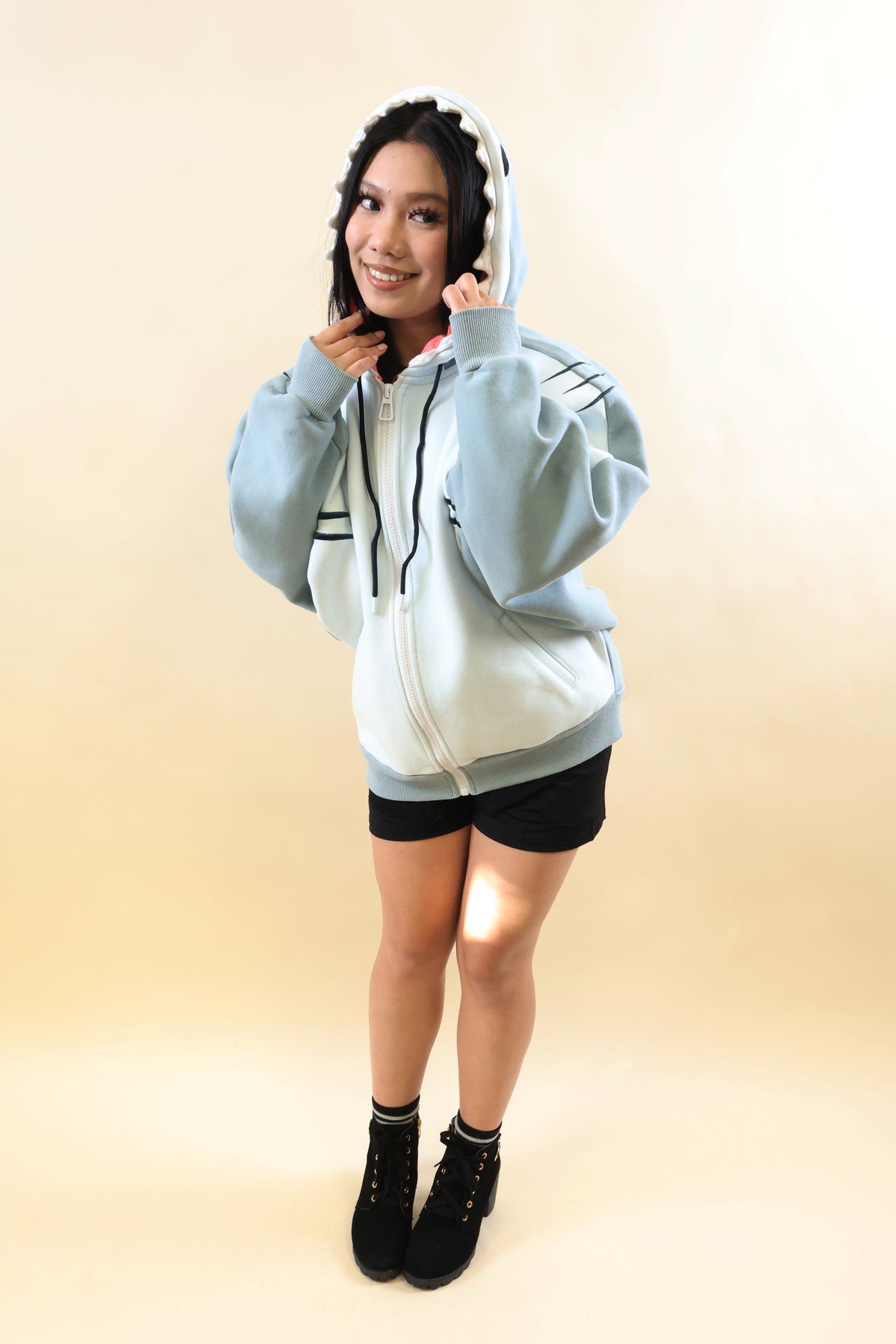 Full-body front view of a smiling model wearing the Nyahallo Shark Hoodie, featuring a light blue and white zip-up design, shark teeth hood, oversized sleeves, embroidered gill details, and front pockets.