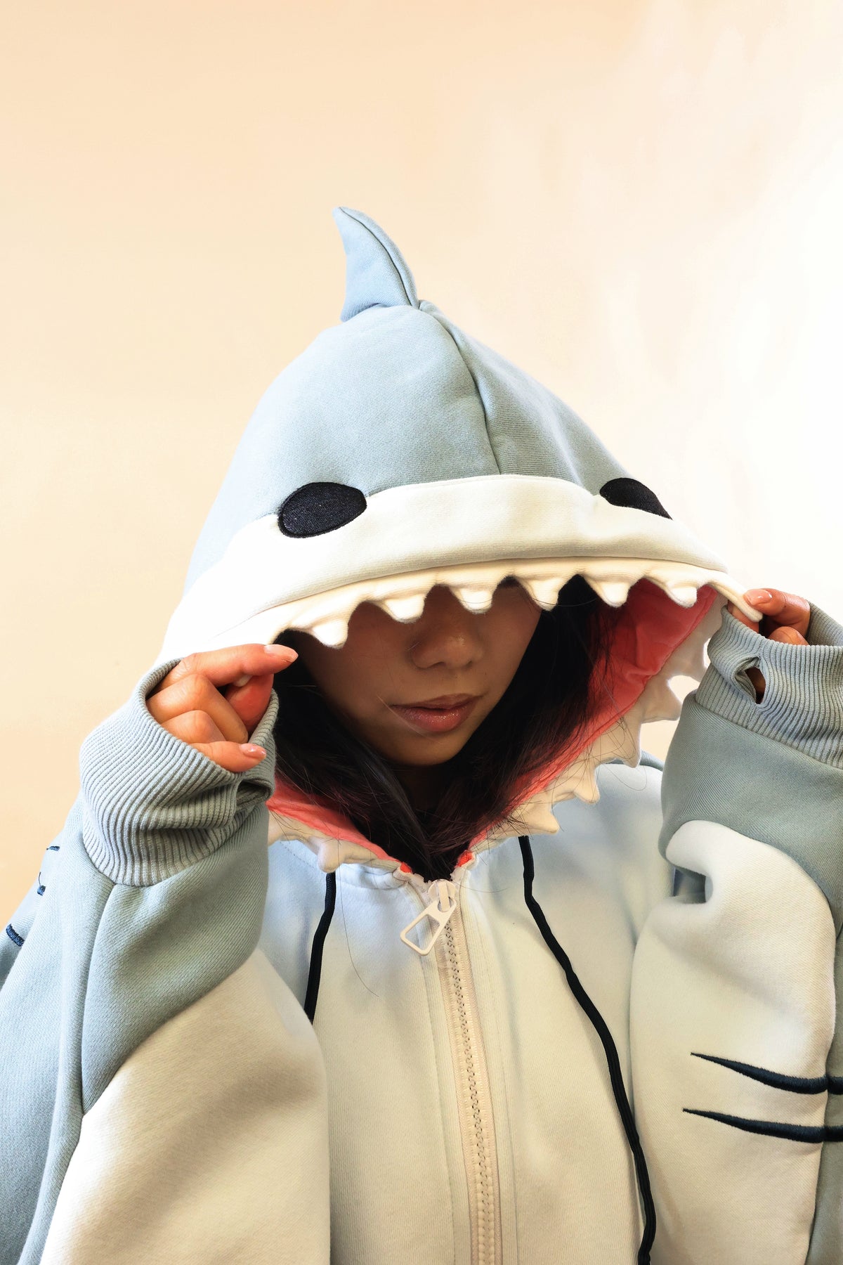 Close-up of a model wearing the Nyahallo Shark Hoodie, holding the light blue shark hood over her eyes, highlighting its dorsal fin, black eyes, white teeth, pink lining, and oversized sleeves.