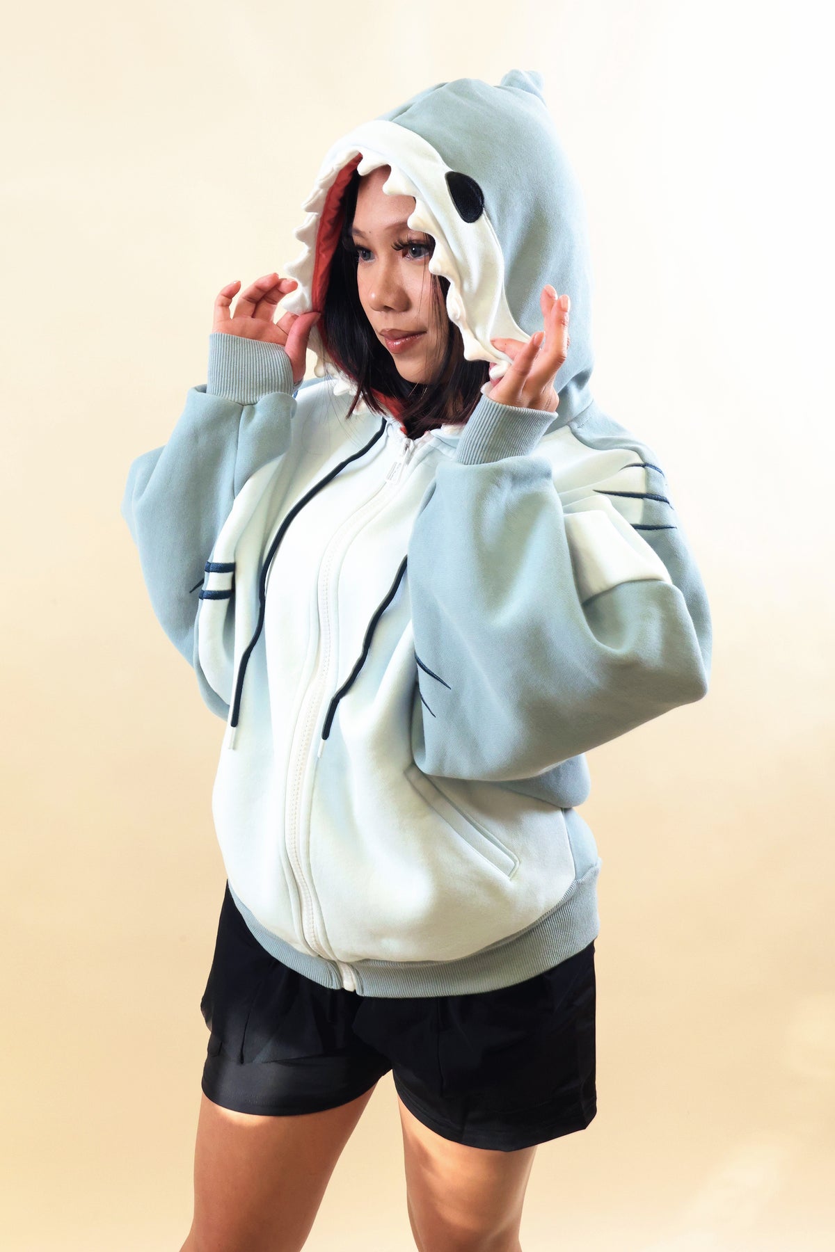 Side view of a model wearing the Nyahallo Shark Hoodie, featuring a light blue and white zip-up design, shark teeth hood with pink lining, embroidered gill details, oversized sleeves, and front pockets.