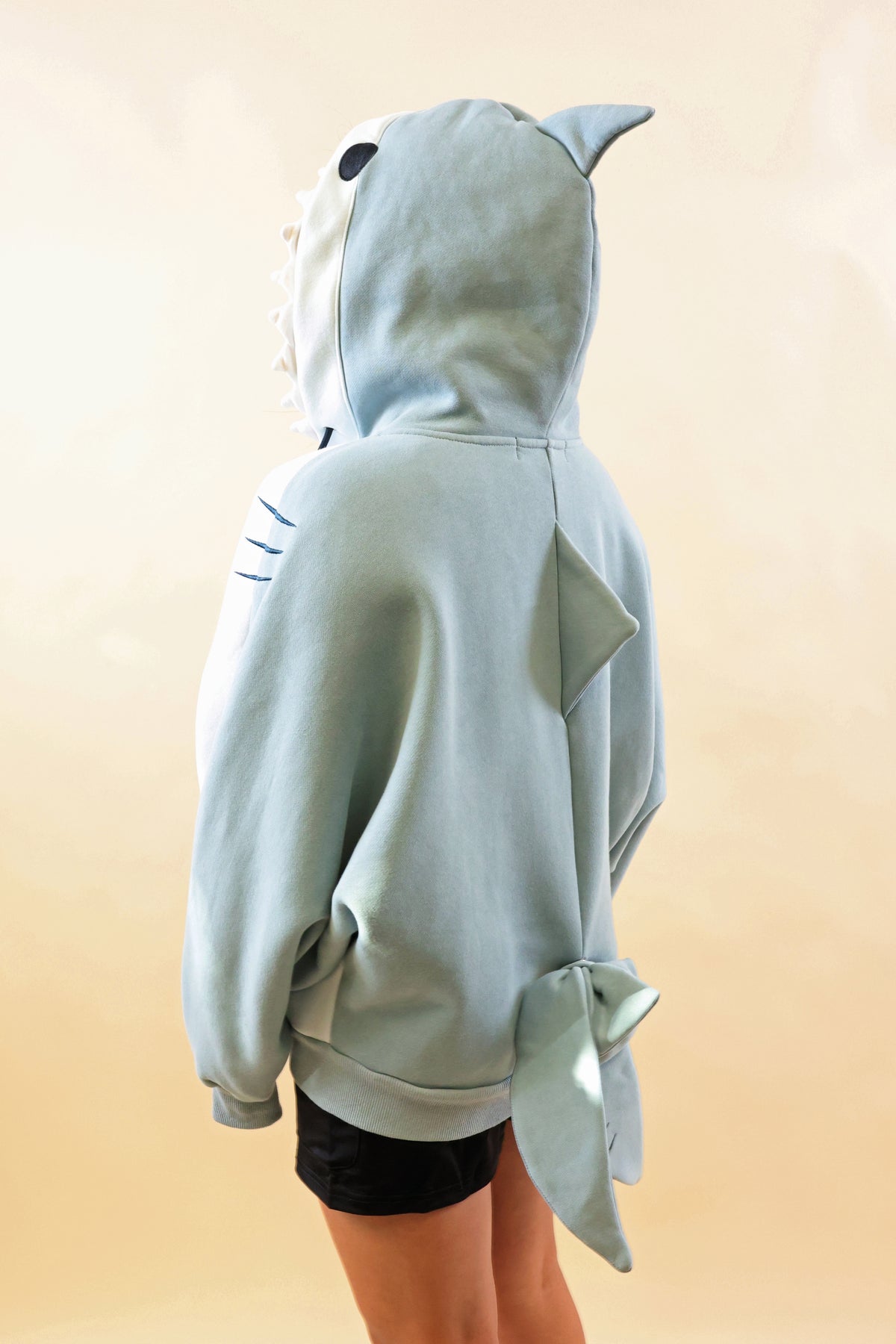Back and side view of a model wearing the Nyahallo Shark Hoodie, featuring a light blue shark hood with dorsal fin, oversized sleeves, a back fin, and a dangling shark tail.