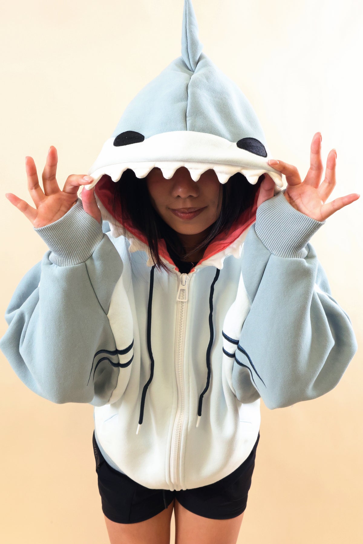 Model wearing the Nyahallo Shark Hoodie, playfully holding the shark hood over her eyes, featuring white teeth, pink lining, dorsal fin, black eyes, and oversized light blue sleeves.
