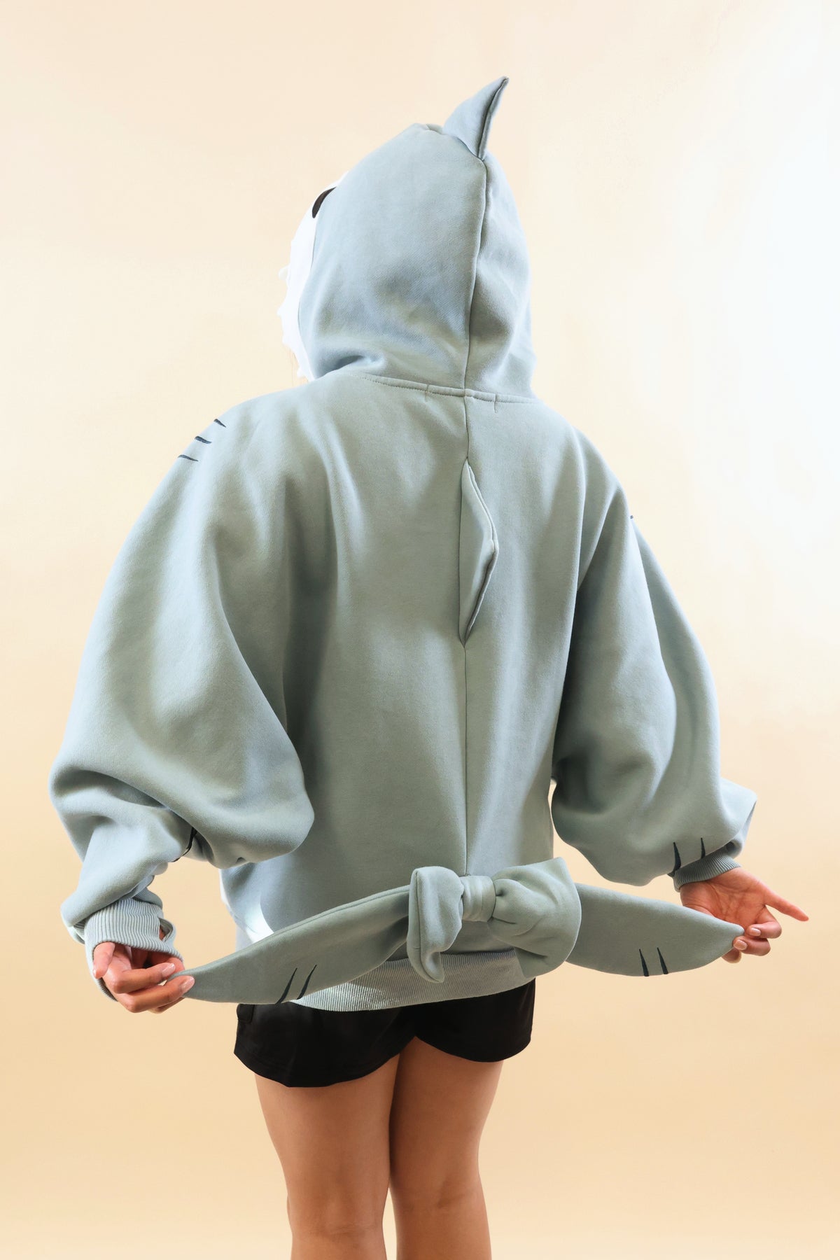 Back view of a model wearing the Nyahallo Shark Hoodie, showcasing its oversized light blue design, dorsal fins, and shark tail held out to display its embroidered details.