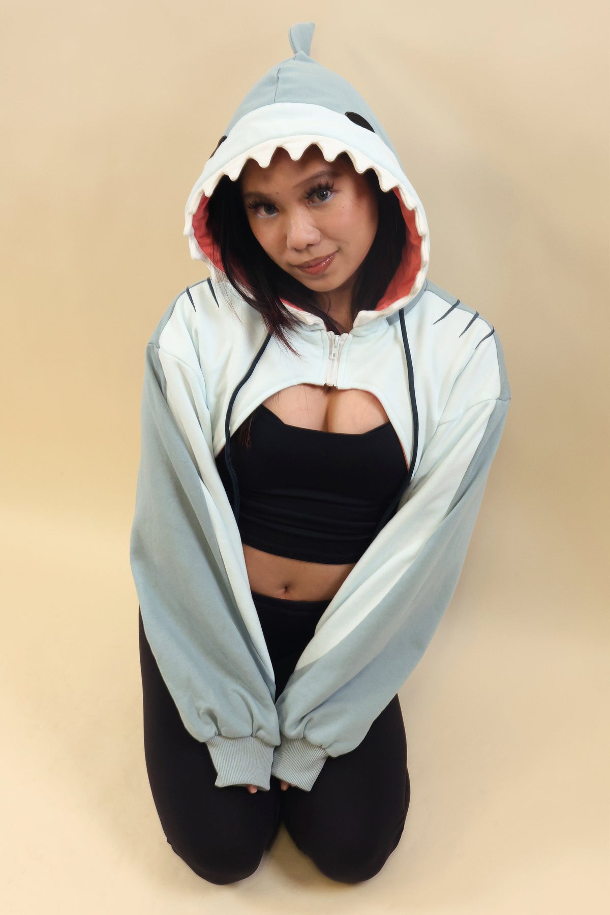 Model wearing the Nyahallo Shark Shrug, featuring a light blue shark hood with white teeth, pink lining, fin detail, and oversized sleeves over a black outfit.