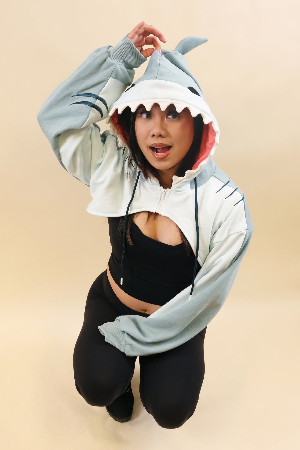 Model kneeling in the Nyahallo Shark Shrug, playfully holding the shark hood fin, featuring white teeth, pink lining, embroidered gill details, and oversized sleeves.