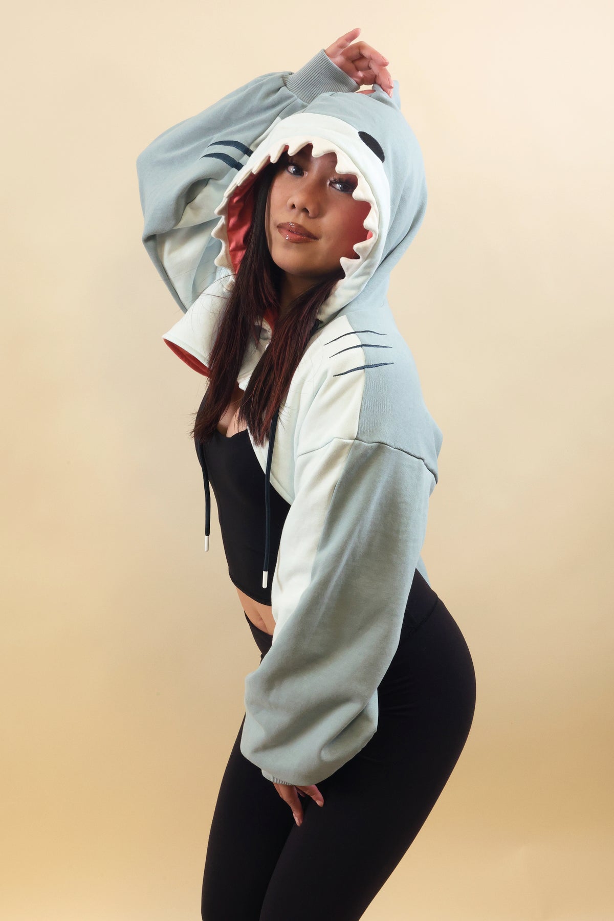 Model posing in the Nyahallo Shark Shrug, featuring a light blue shark hood with white teeth, pink lining, embroidered gill details, and oversized sleeves.