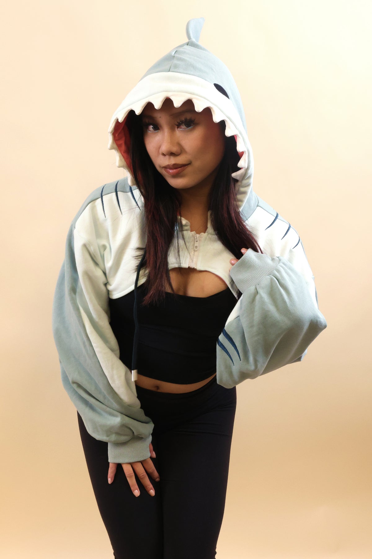 Model wearing the Nyahallo Shark Shrug, featuring a light blue shark hood with white teeth, pink lining, dorsal fin, embroidered gill details, and oversized sleeves over a black outfit.