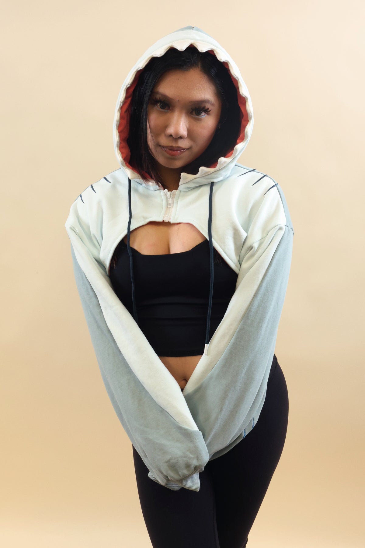 Front view of a model wearing the Nyahallo Shark Shrug, featuring a light blue and white cropped design, shark teeth hood with pink lining, oversized sleeves, and adjustable drawstrings.