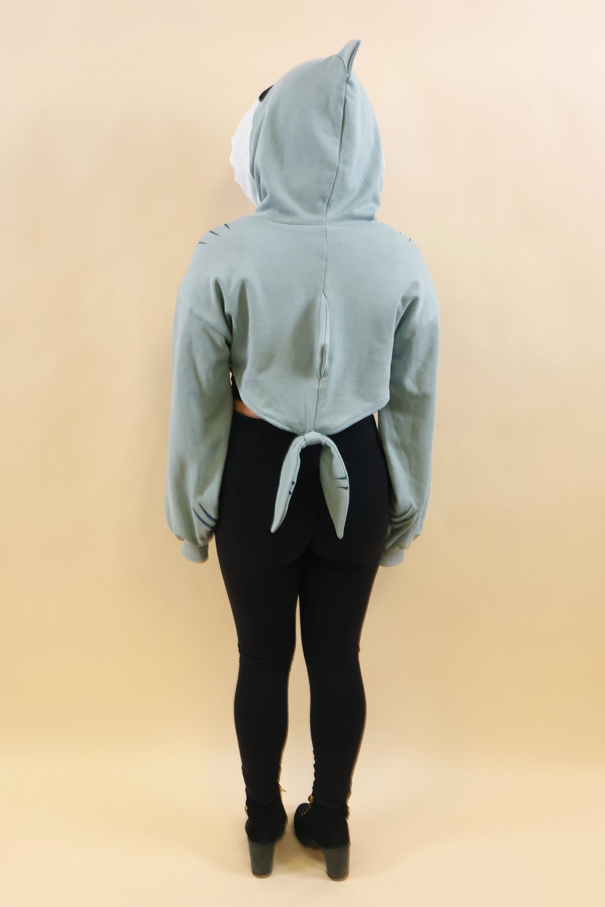 Full back view of a model wearing the Nyahallo Shark Shrug, featuring a light blue cropped design, shark hood with dorsal fin, oversized sleeves, back fin, and dangling shark tail.