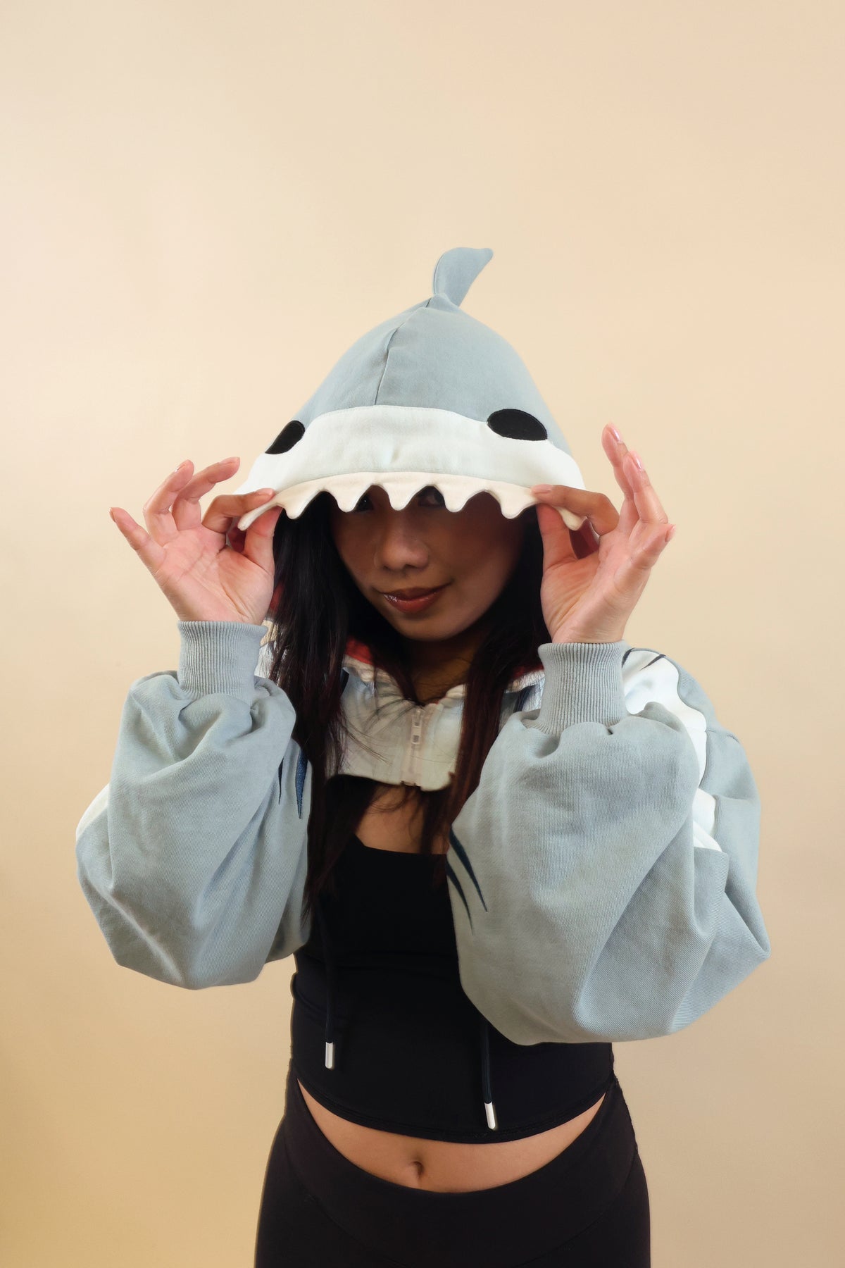 Model wearing the Nyahallo Shark Shrug, holding the shark hood over her eyes to highlight its white teeth, black eyes, dorsal fin, and oversized light blue sleeves.