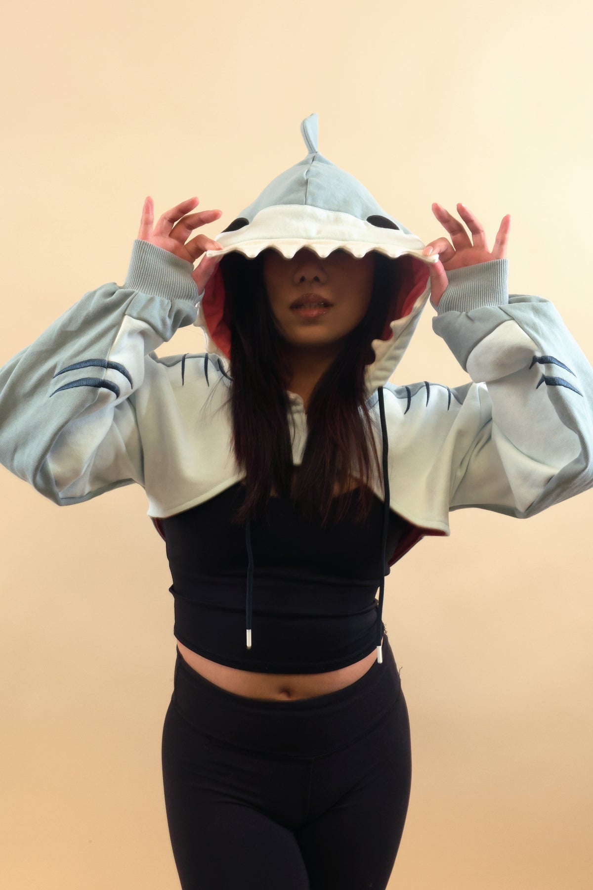 Model wearing the Nyahallo Shark Shrug, holding the shark hood over her eyes, featuring white teeth, pink lining, dorsal fin, embroidered gill details, and oversized sleeves.