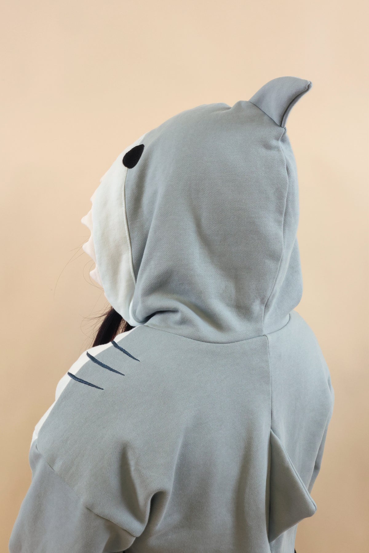 Close-up back and side view of the Nyahallo Shark Shrug, highlighting its light blue shark hood with dorsal fin, black eye, white teeth, embroidered gill details, and back fin.
