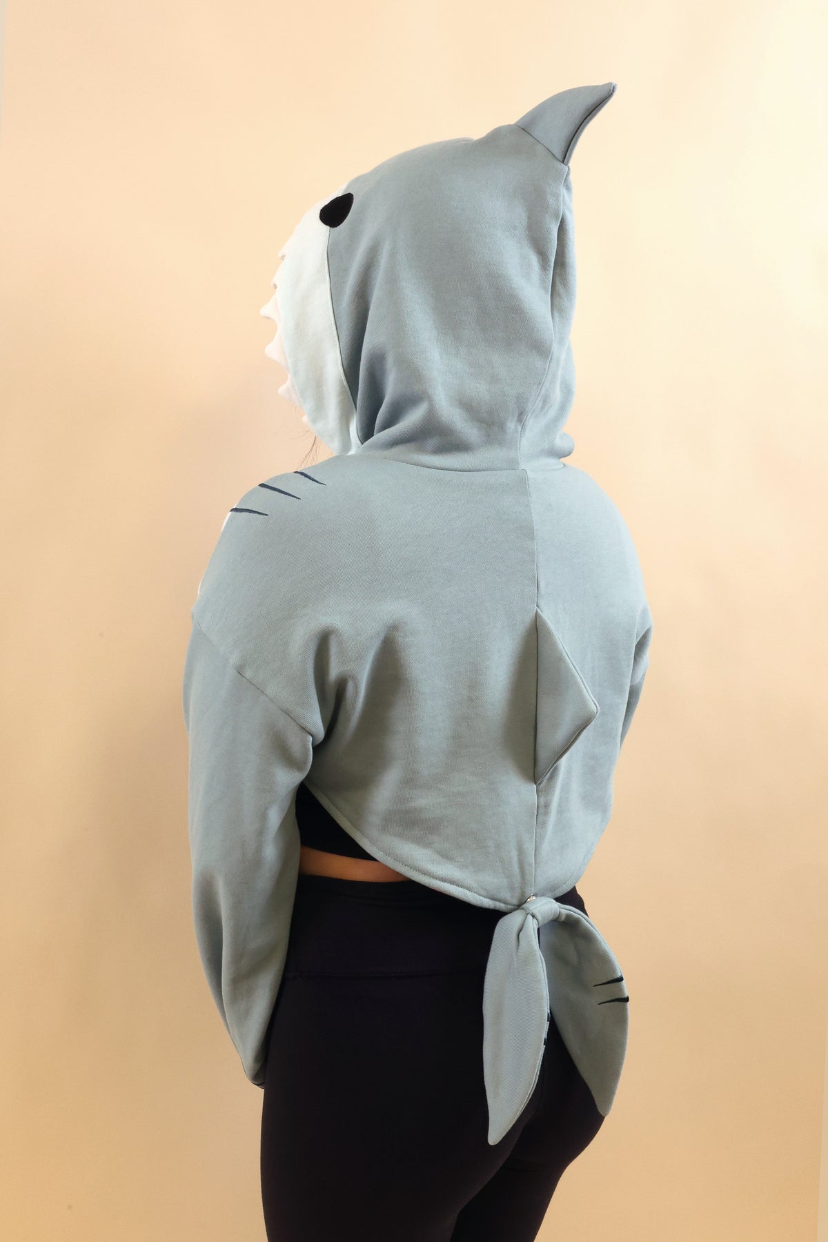 Back view of a model wearing the Nyahallo Shark Shrug, featuring a light blue shark hood with dorsal fin, oversized sleeves, and a shark tail detail at the back.