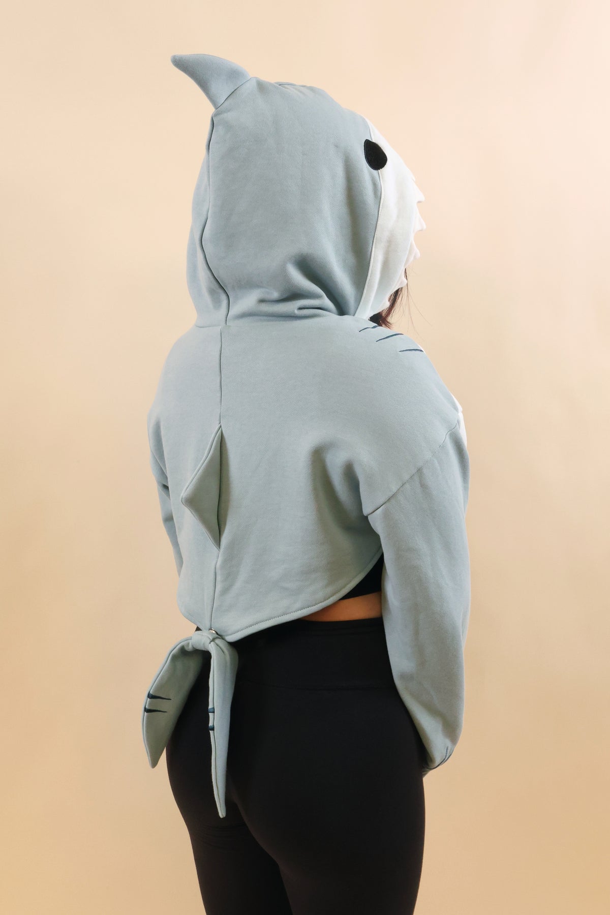 Rear three-quarter view of a model wearing the Nyahallo Shark Shrug, featuring a light blue shark hood with dorsal fin, a back fin, oversized sleeves, and a dangling shark tail.